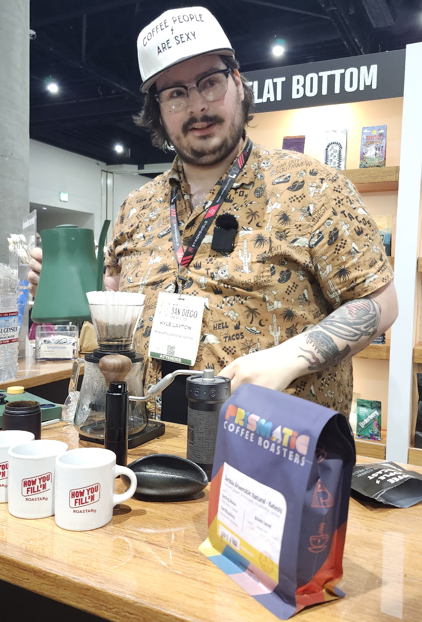 In a packaging display booth in a convention hall, a barista brews a pourover coffee. The table in front of him is filled with coffee mugs, bags, and brewing equipment. He's looking at the camera with a quizzical expression.
