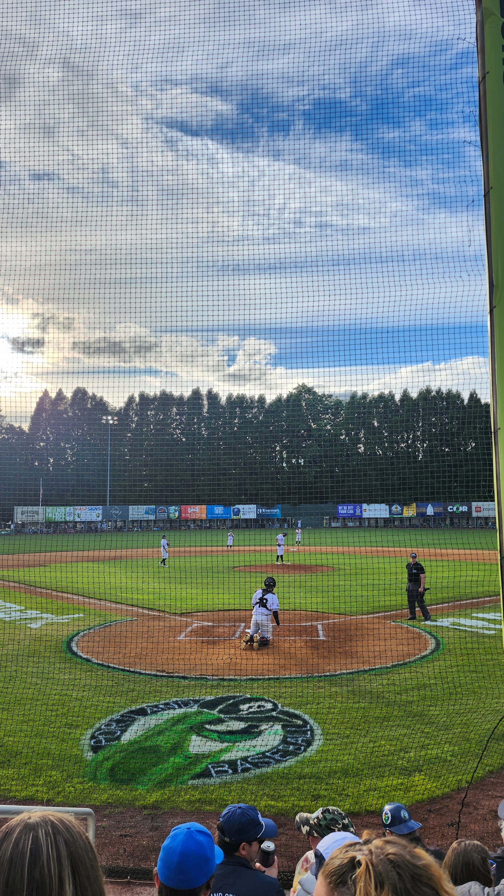 A baseball pitcher prepares to throw a warm-up pitch to the catcher. Photo taken from behind home plate. Some fans holding beverages are in the foreground.