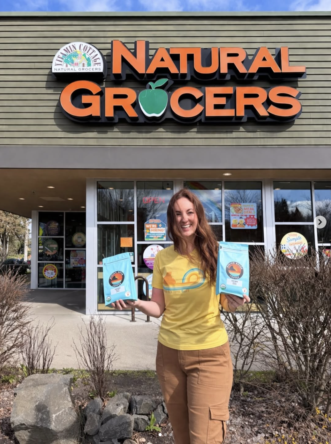 Long brunette hair blows across the face of a smiling women holding up two bright blue coffee bags to the camera outside in front of a grocery store entrance.