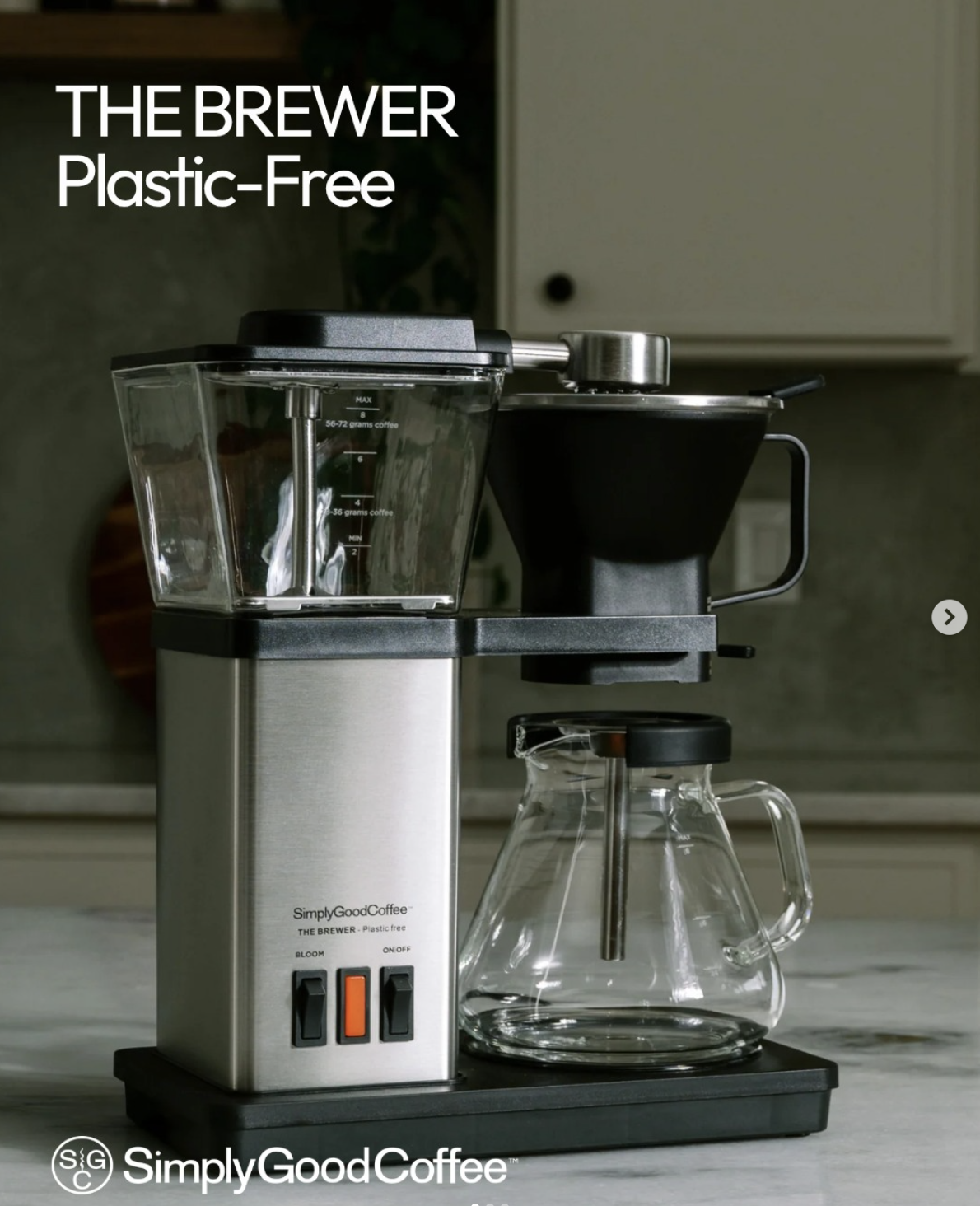 A plastic-free countertop coffee brewer sits on a white and black countertop. The brewer features a silver tower with two black buttons of on/off and bloom features and an orange light indicator for brewing. A clear-glass water tank sits on top of the tower and a dispersal arm stretches out over a black v60 inspired brew basket. Below the basket, a glass pot with black lid waits for coffee to drip down.
