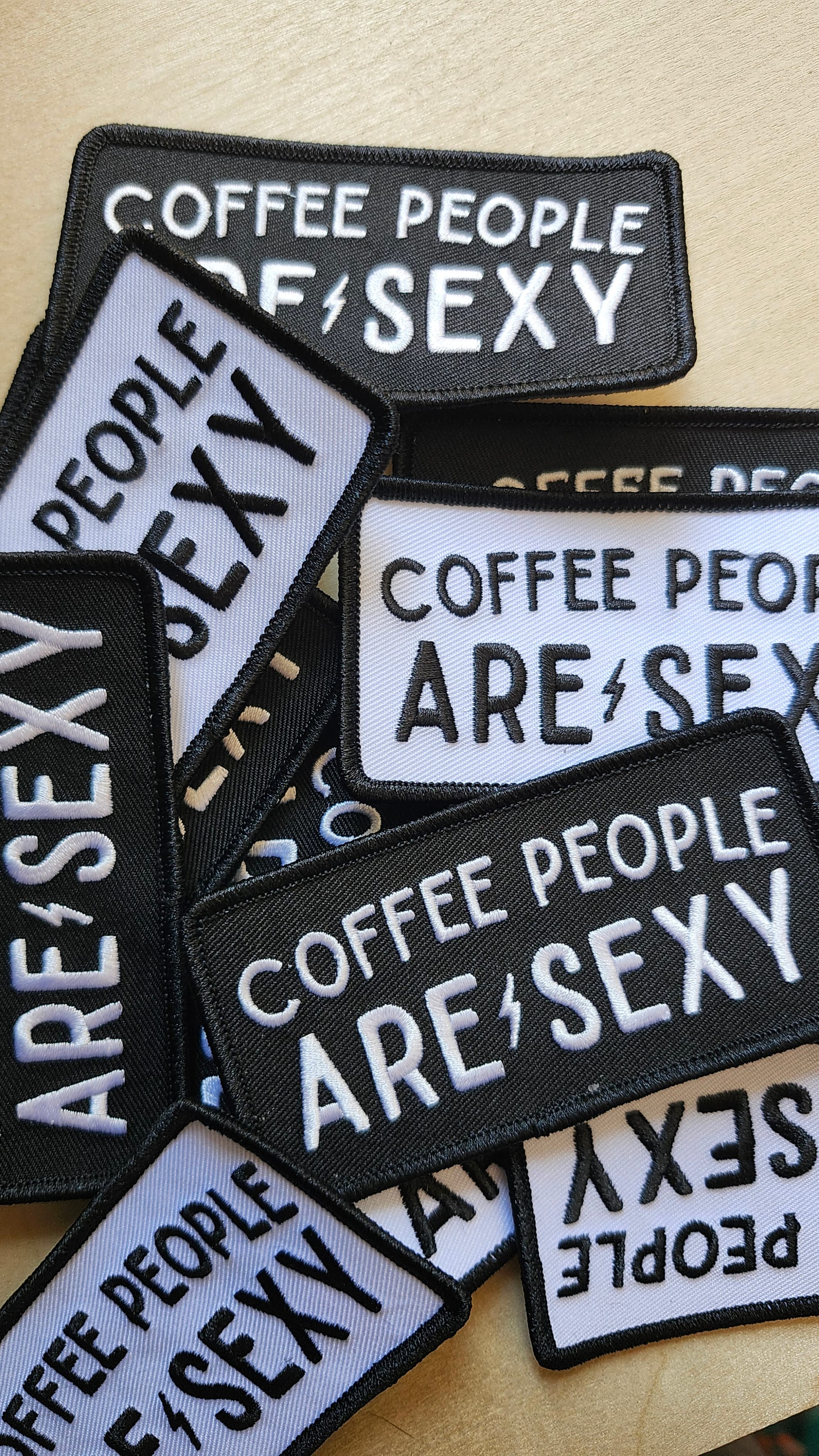 A close-up photo of horizontal black and white or white and black patches that say Coffee People Are Sexy with a small lightning bolt between "are" and "sexy."