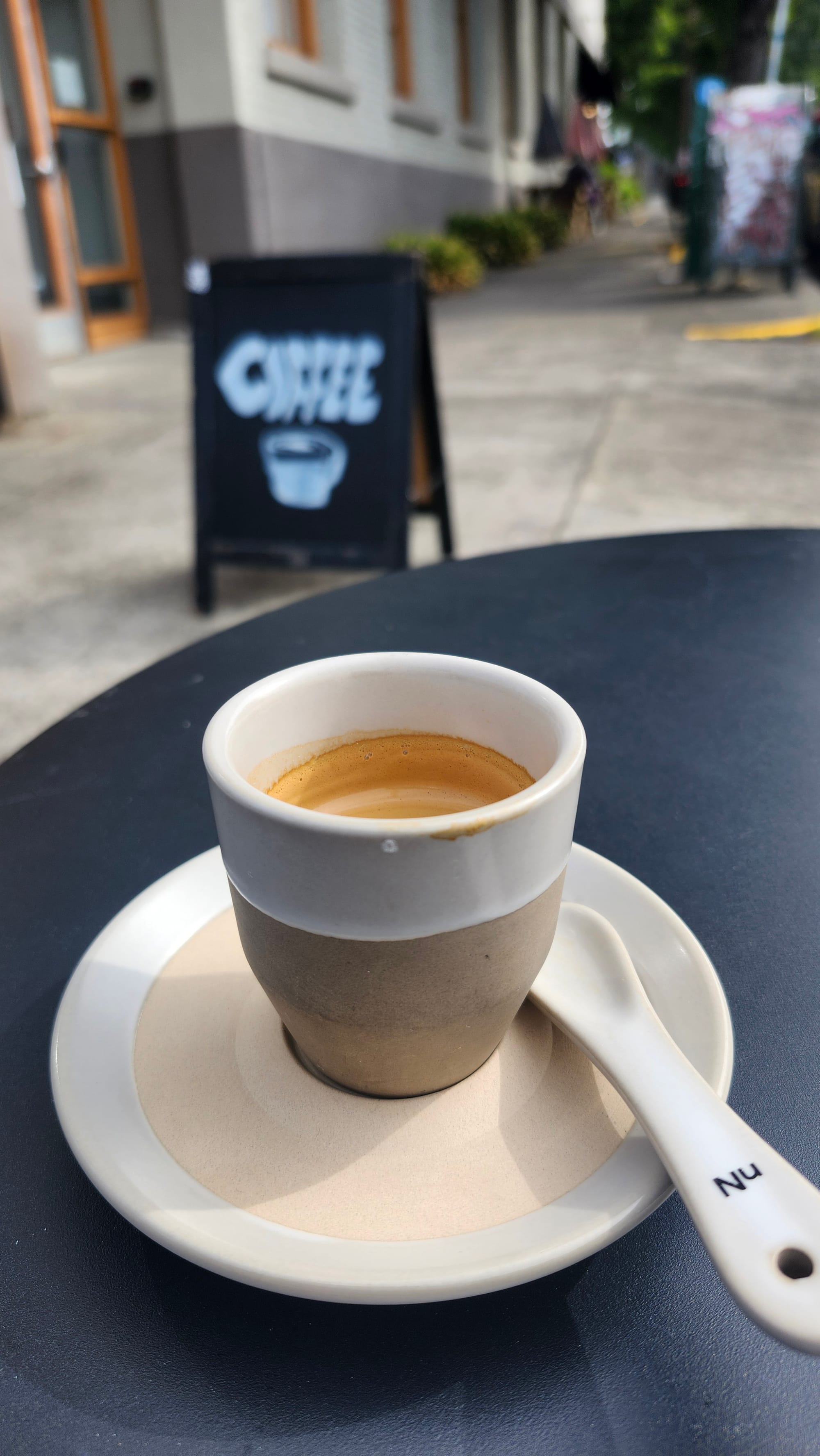 A close up on a two-tone cream espresso cup with a natural brown clay base on a matching saucer with a clay spoon. The remains of espresso coat the inside of the glass and a hand-chalked A-frame sidewalk sign that says "Coffee" with a drawing of a coffee cup and saucer is blurred on the sidewalk in the background.