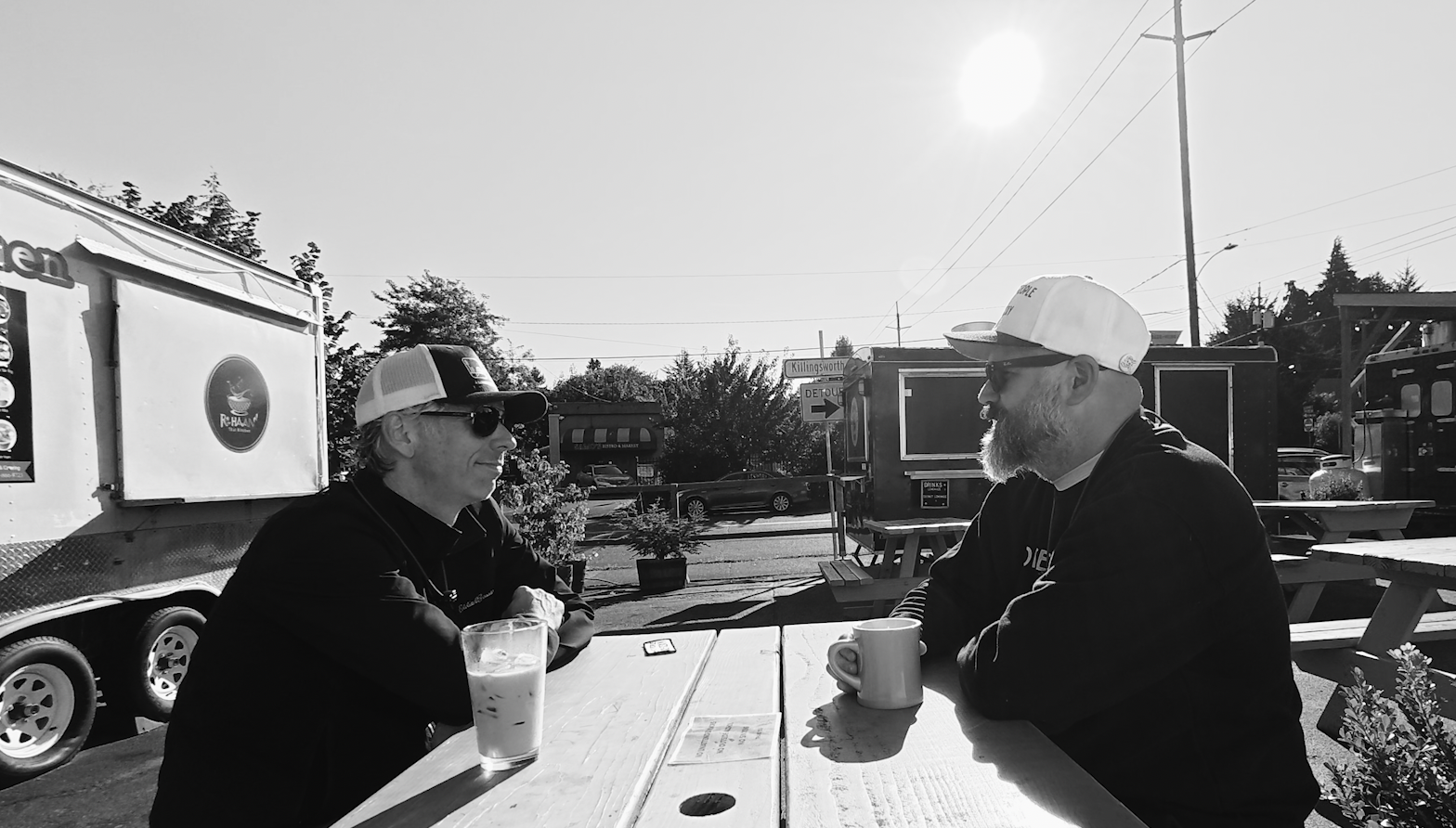 A black-and-white image of two men in black sweatshirts, sunglasses, and baseball caps sitting across from each other at picnic table in a food truck court. Coffee mugs are on the table in front of them.