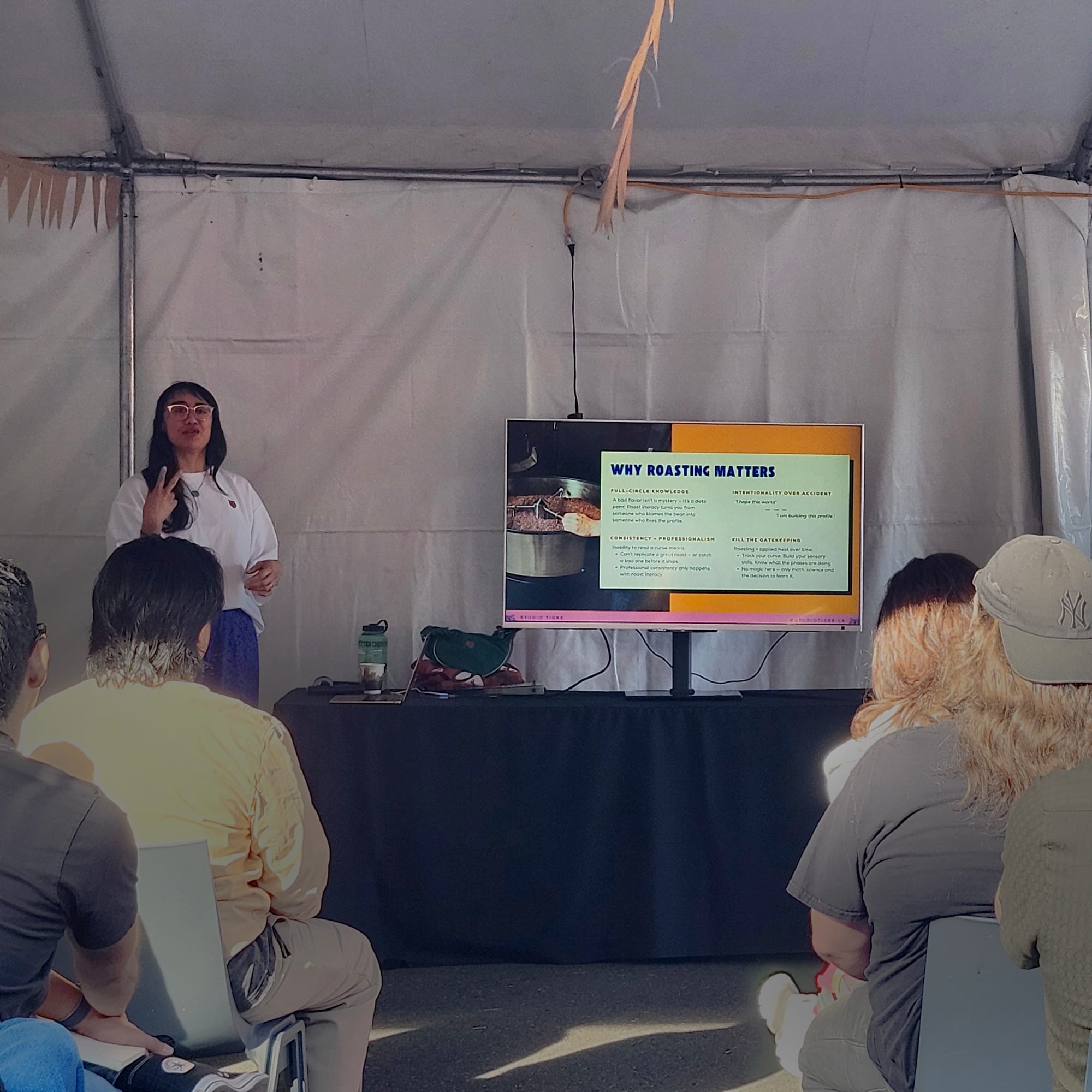 A woman holds up two fingers while speaking to a group of attendees at a roasting education seminar in an outdoor tent with a presentation screen with written information featured prominently at a head table at the front of a group