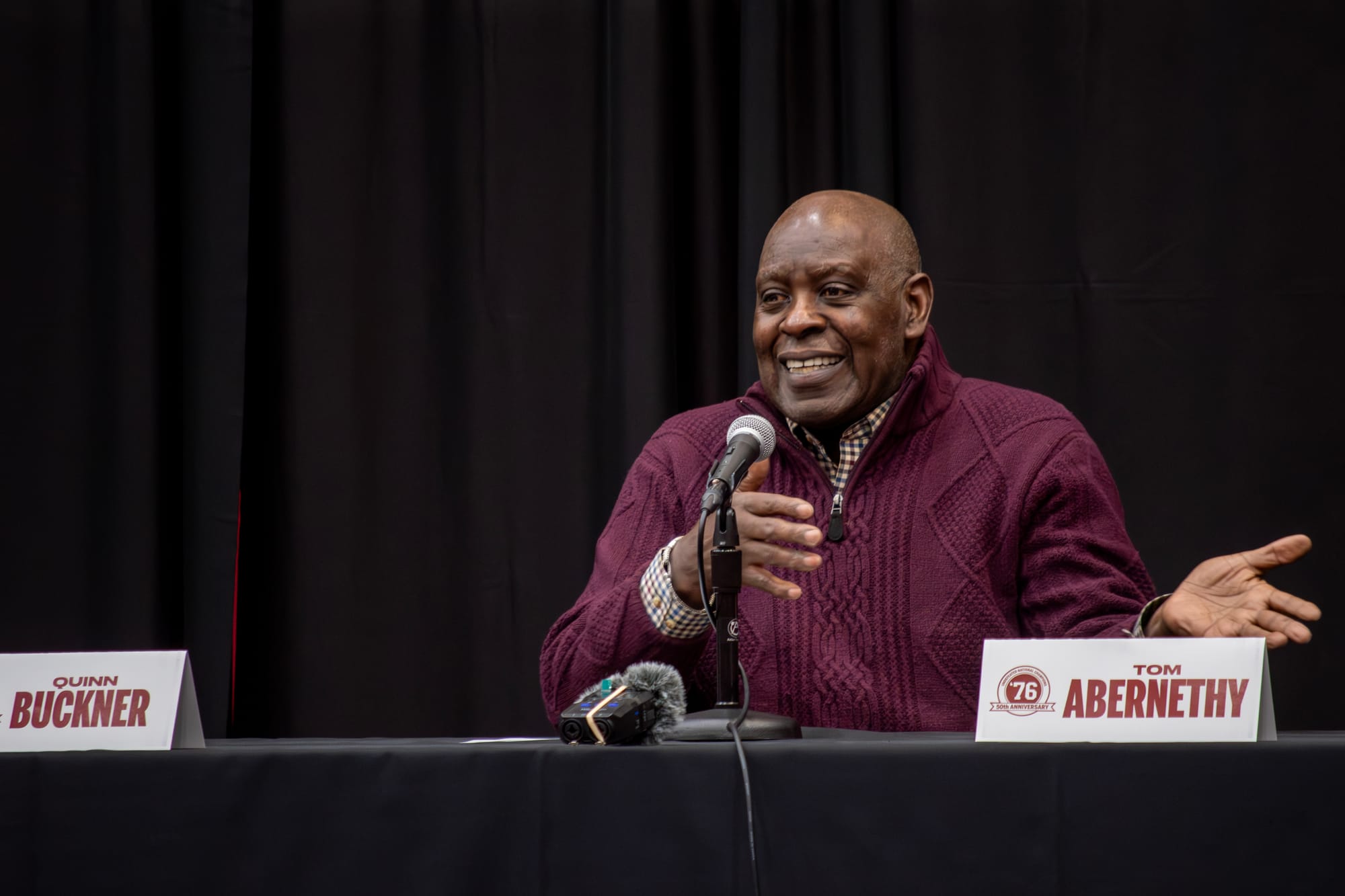 Quinn Buckner, a 71-year-old black man, wears a purple quart-zip sweater while sitting behind a microphone at a black table 