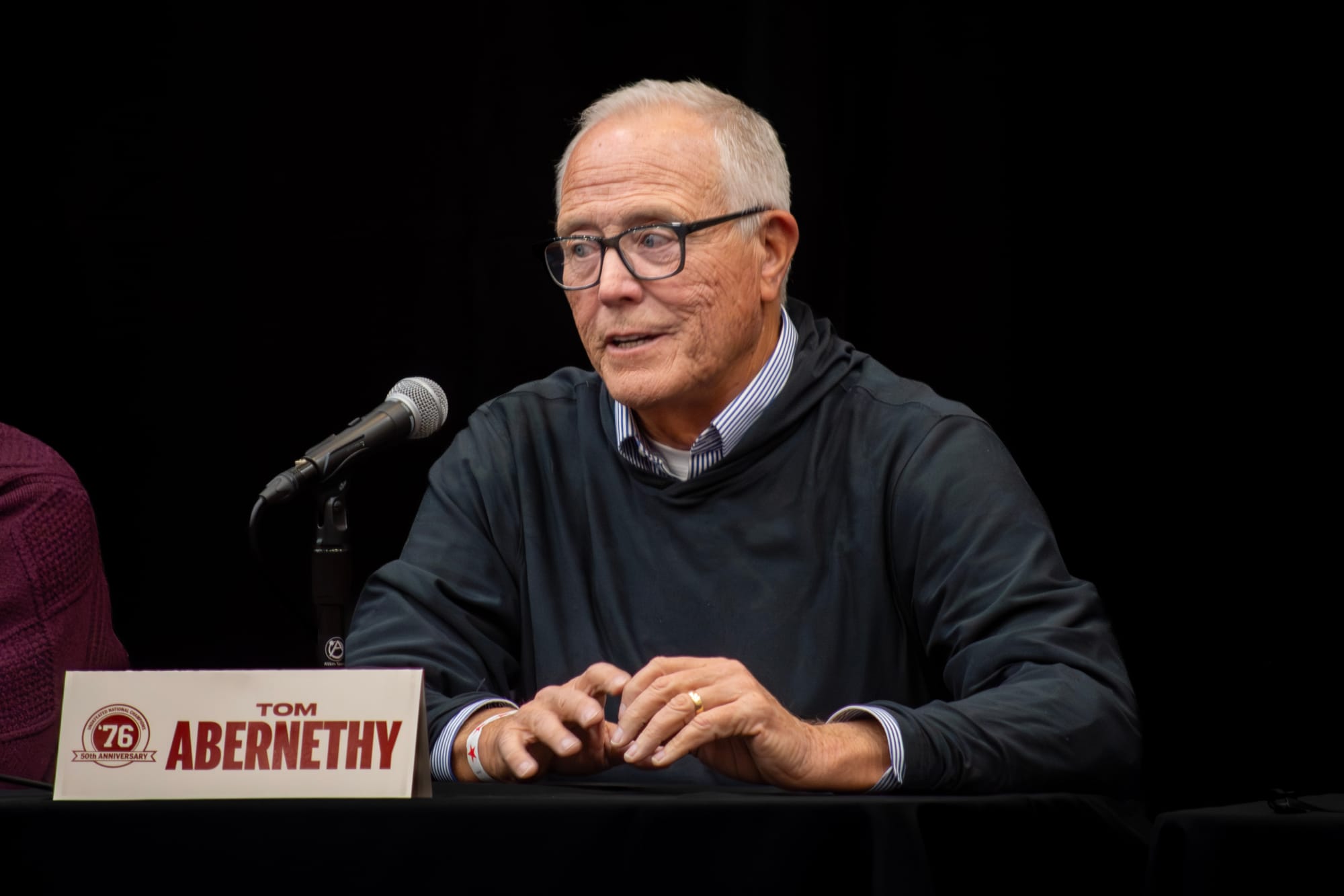 Tim Abernethy, a 71-year-old white man wearing a black shirt and glasses, speaks into a microphone