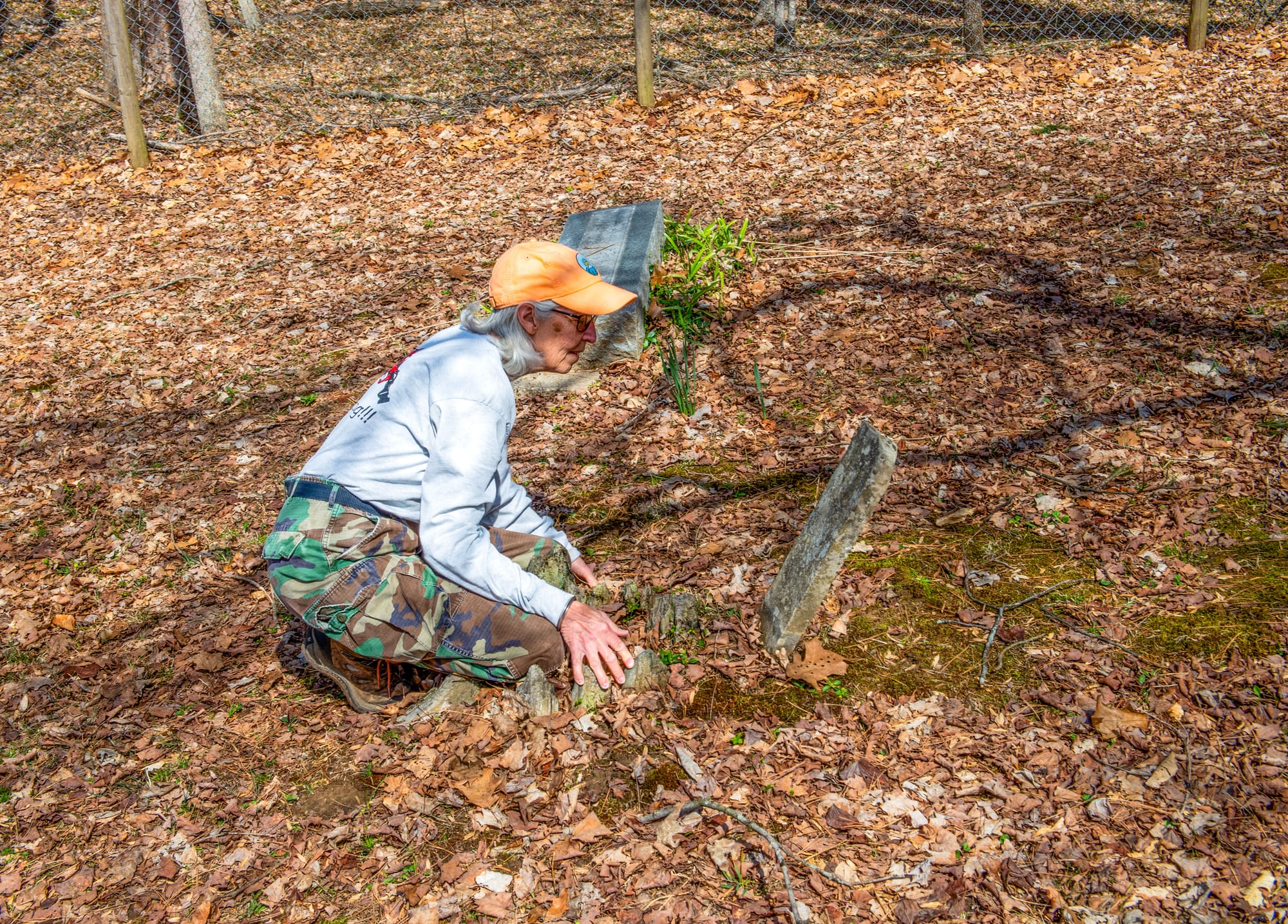 an elderly woman wearing an orange hat, glasses, a grey long-sleeved shirt, and camouflage pants kneels in front of an old headstone 