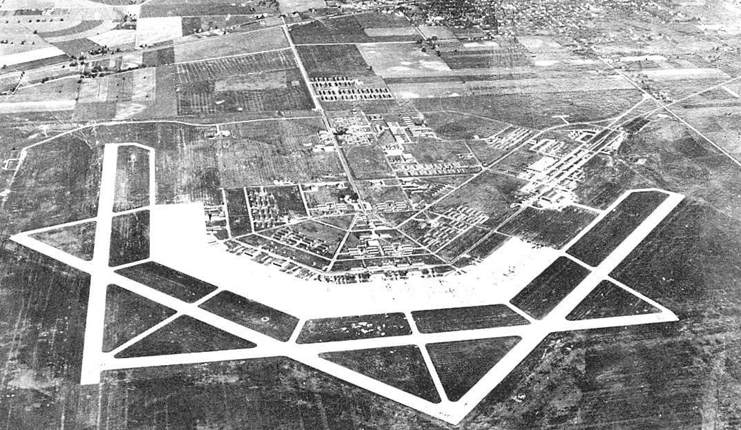 a black and white aerial photograph of an Army airfield base amidst fields and urban development
