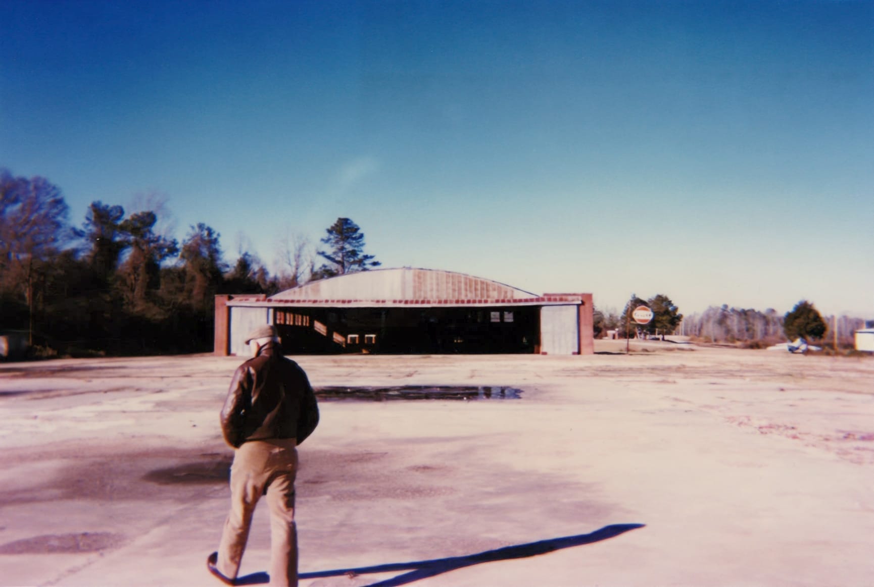 a man wearing a flat cap, a brown leather jacket, and brown pants walks with hands in pockets toward a rusted airfield hangar