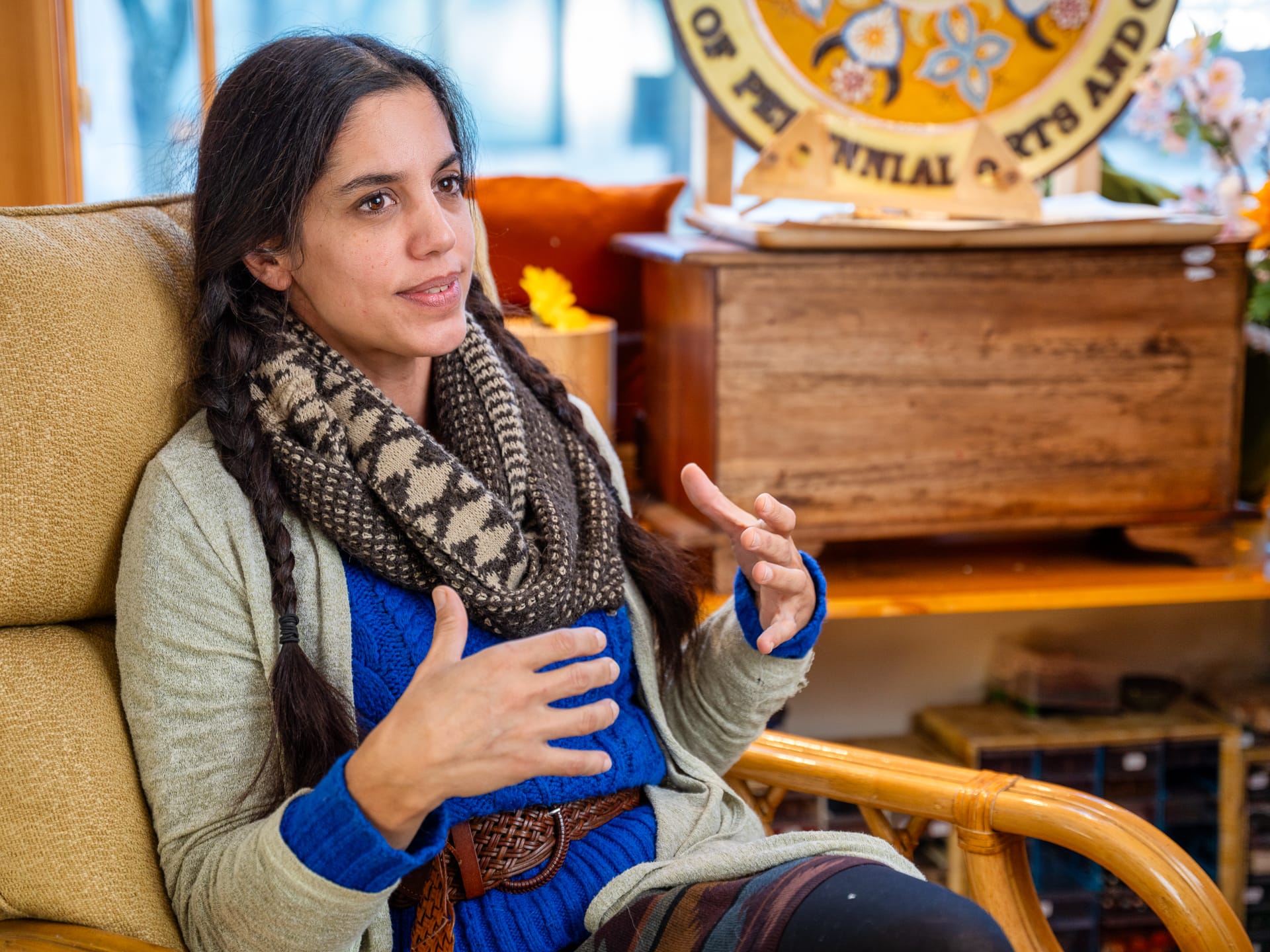 Heidi Fitzgerald wears braids in her dark brown hair, a multi-toned brown scarf, a light brown cardigan, and a blue knit sweater. She sits 