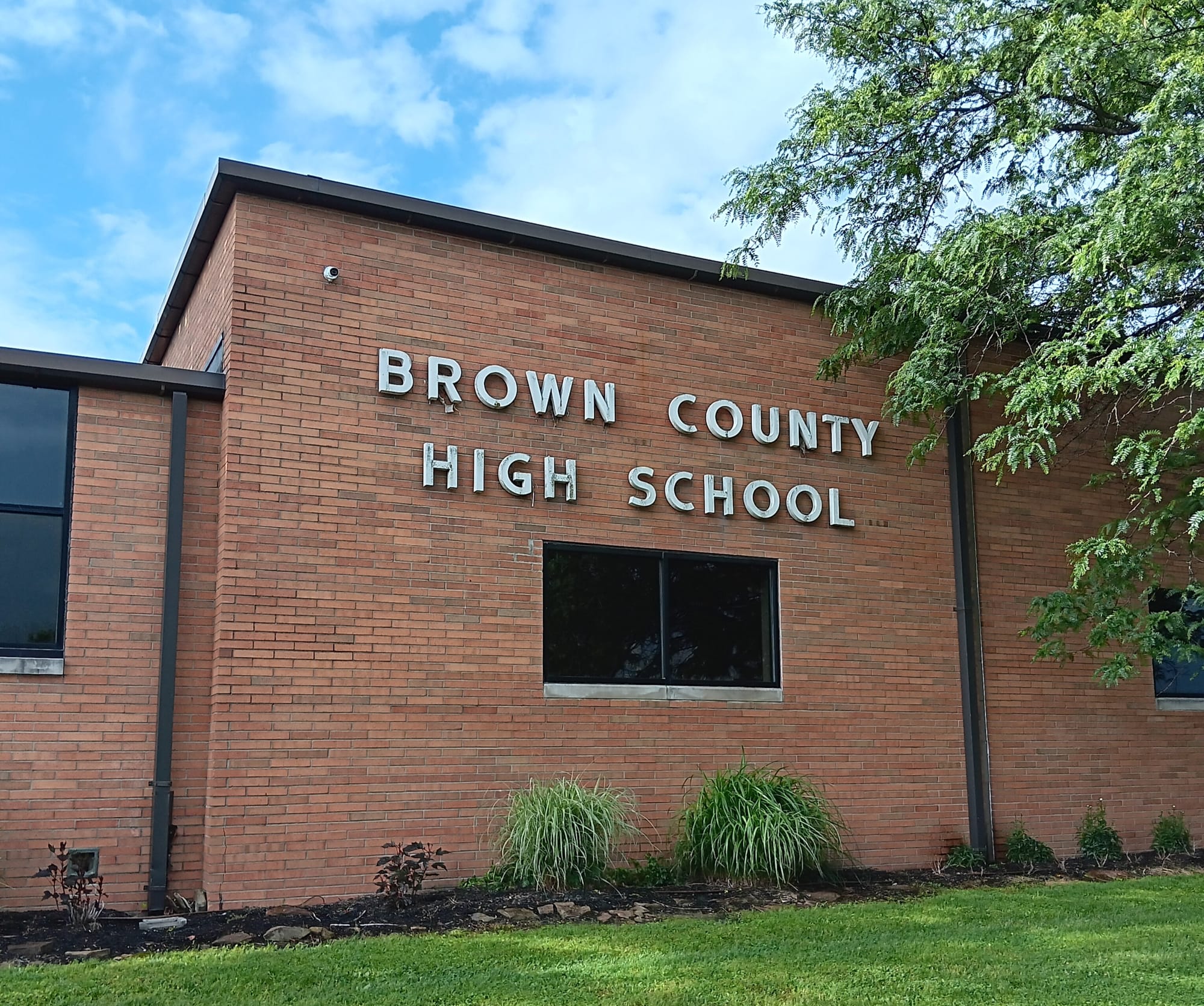 a squarish, red brick exterior of the one-story Brown County High School building. A tree overhangs the right side of the image; some ornamental grasses and small shrubs fill the landscaping in front of the wall. A blue sky with cottony white clouds shines brightly behind the building