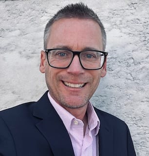 a white man with gray hair, black-rimmed glasses, and a blue blazer