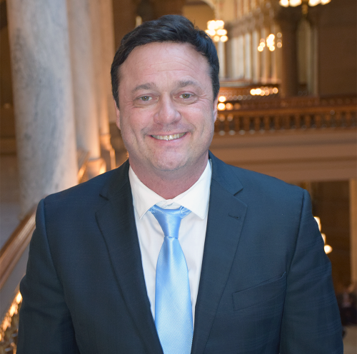 a middle-aged white man wearing a blue blazer, white dress shirt, and sky blue tie, pictured inside an ornate government building