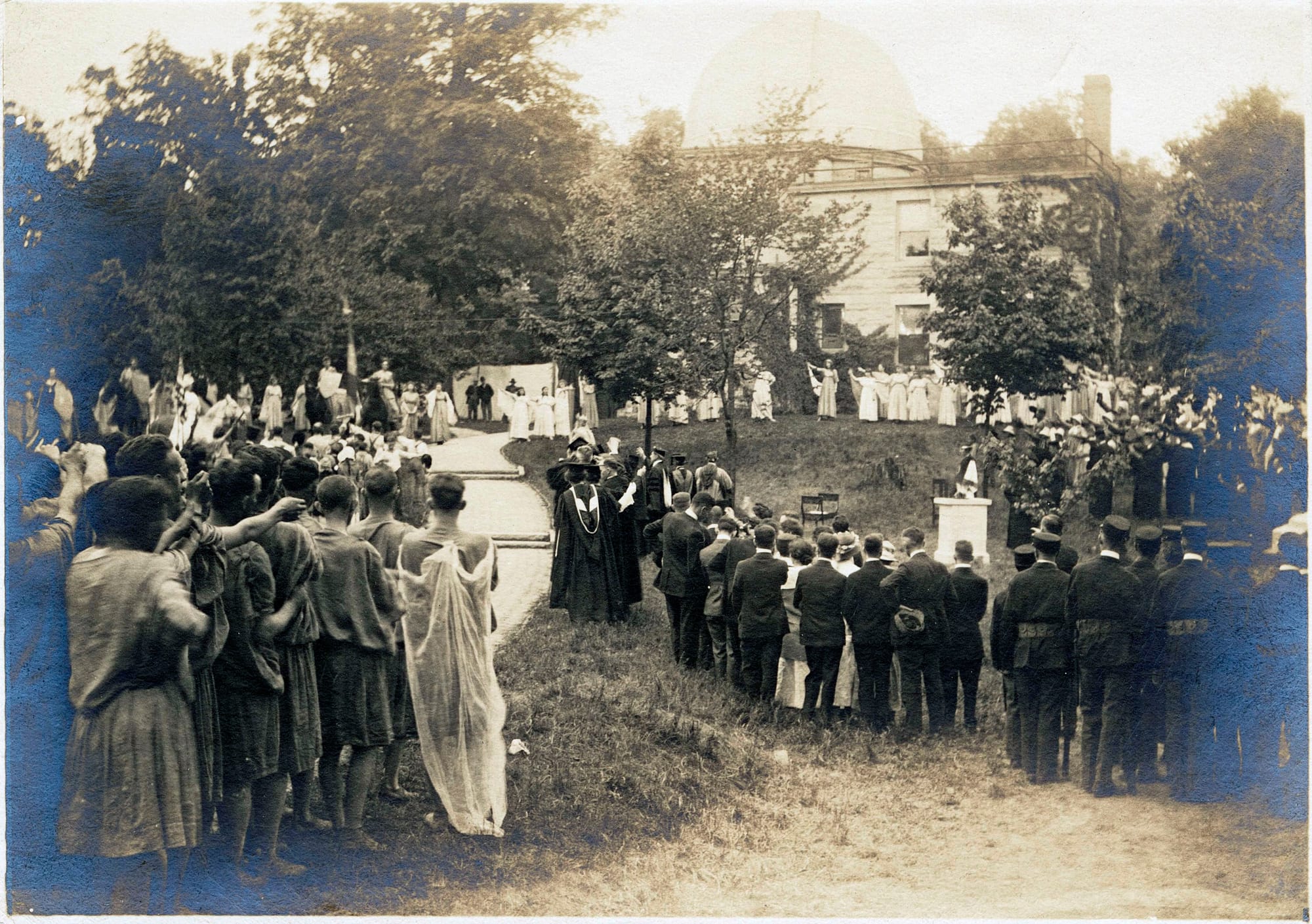 a sepia-toned photograph of costumed performers gathering in a choreographed procession on a grassy slope beneath a limestone observatory building.