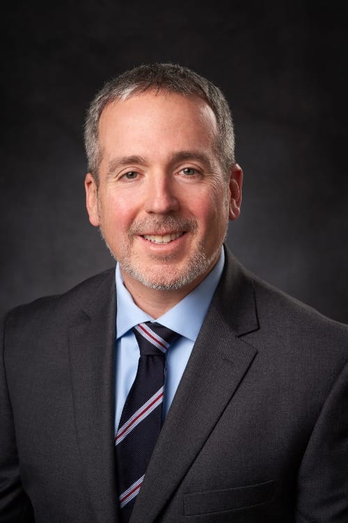 a middle-aged white man in a gray blazer and sky blue button up shirt with a blue, red, and white tie 