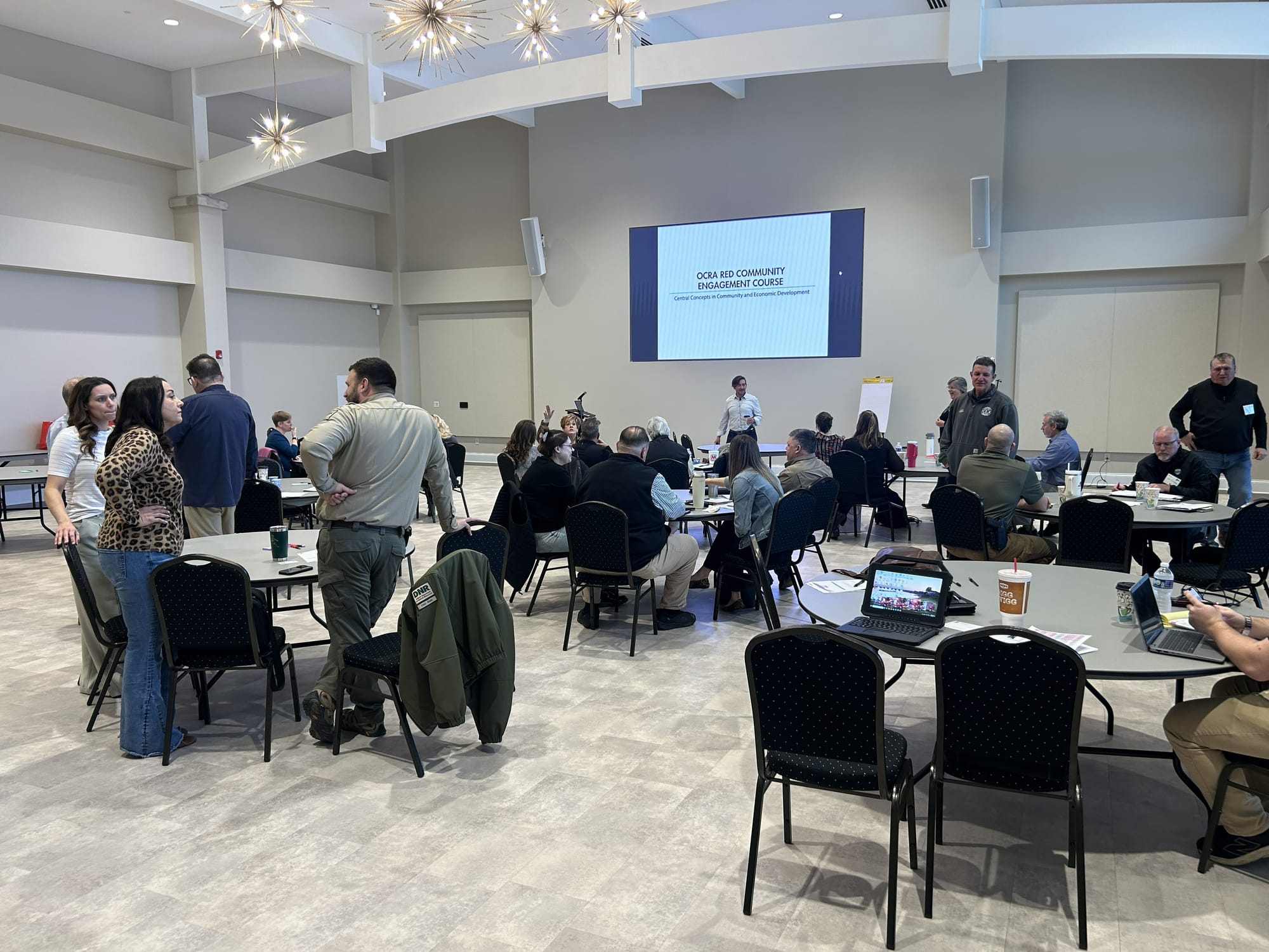 round tables and padded black chairs fill a large event space room as program participants mingle