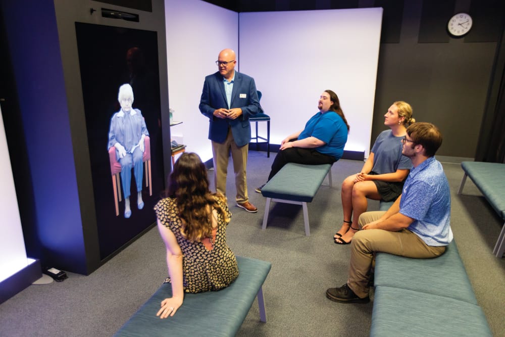 four individuals sit on benches in a room; one museum guide in a blue blazer stands beside a visual display screen depicting an old woman in a chair