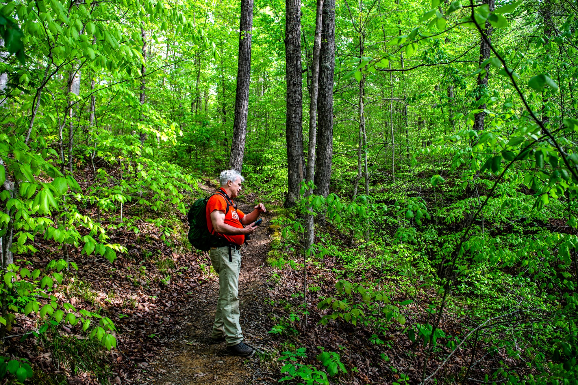 grey-haired, orange-shirt-wearing Jeff Stant stands and gestures on a steep trail leading up a wooded hillside