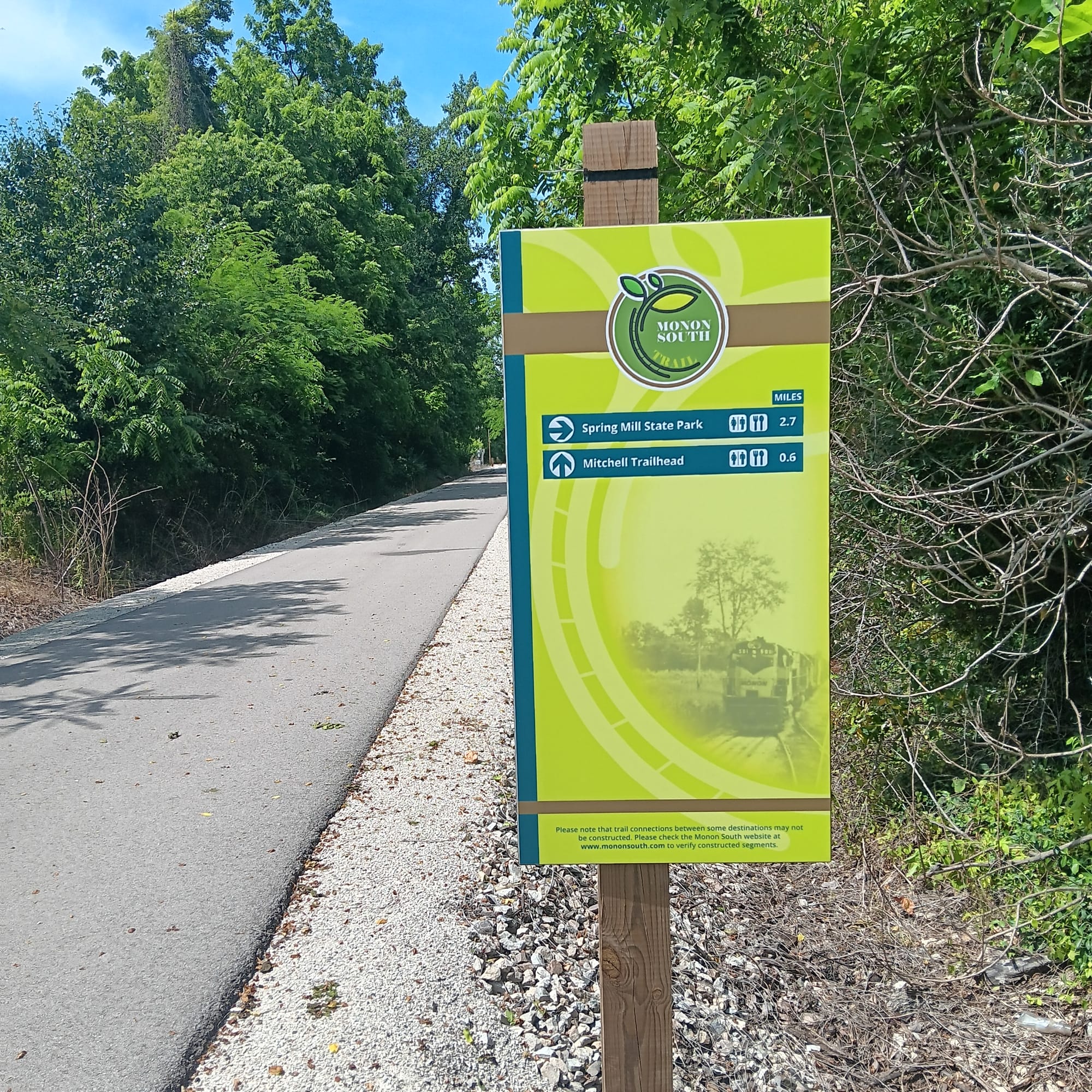 a flat paved trail runs over the gravel of what was once a railbed. A signboard indicates the name of the trail and nearby destinations (Spring Mill State Park; Mitchell Trailhead)