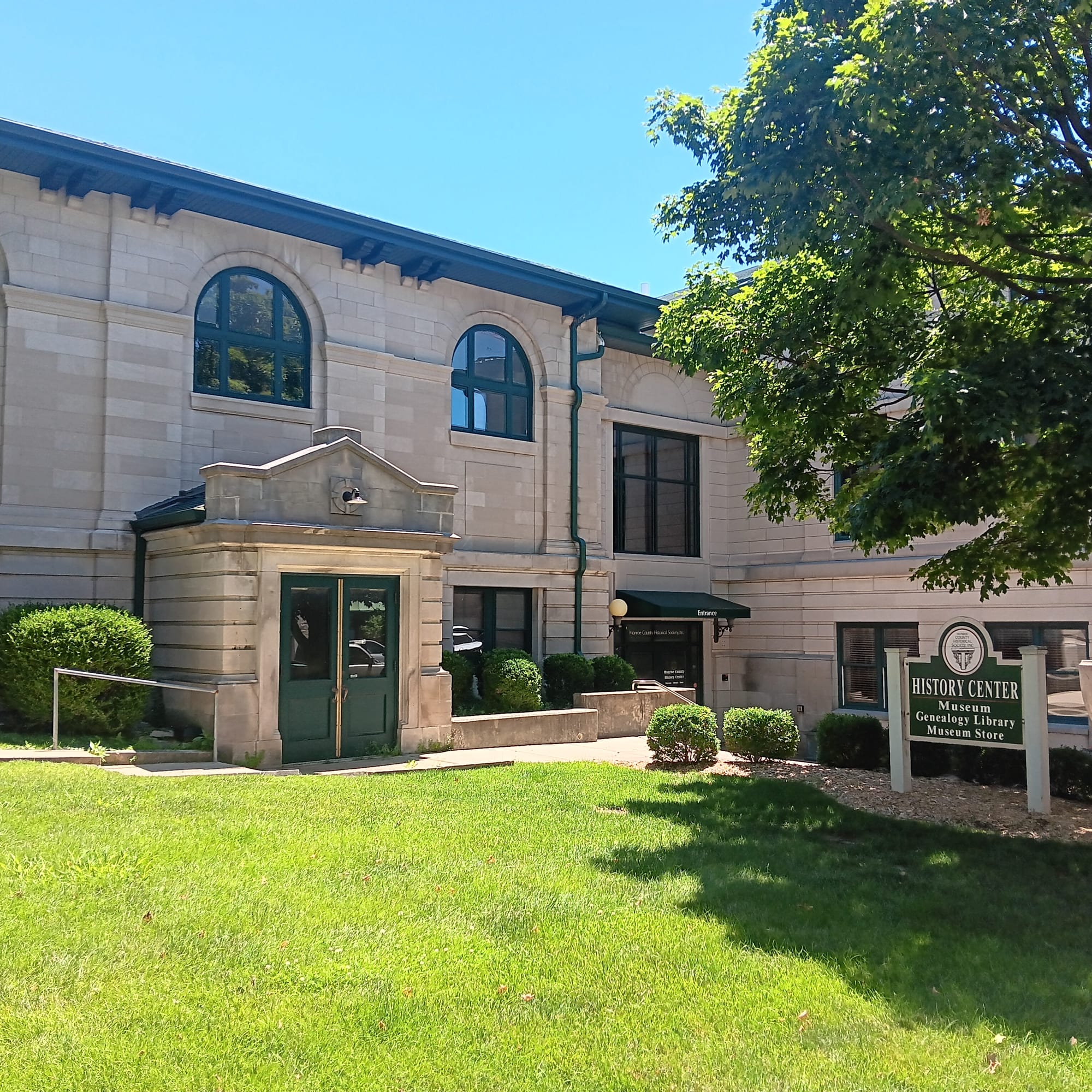 a two story limestone building, grassy lawn, maple tree, and small shrubs. A wooden sign painted white and green says "HISTORY CENTER" Museum, Genealogy Library, Museum Store