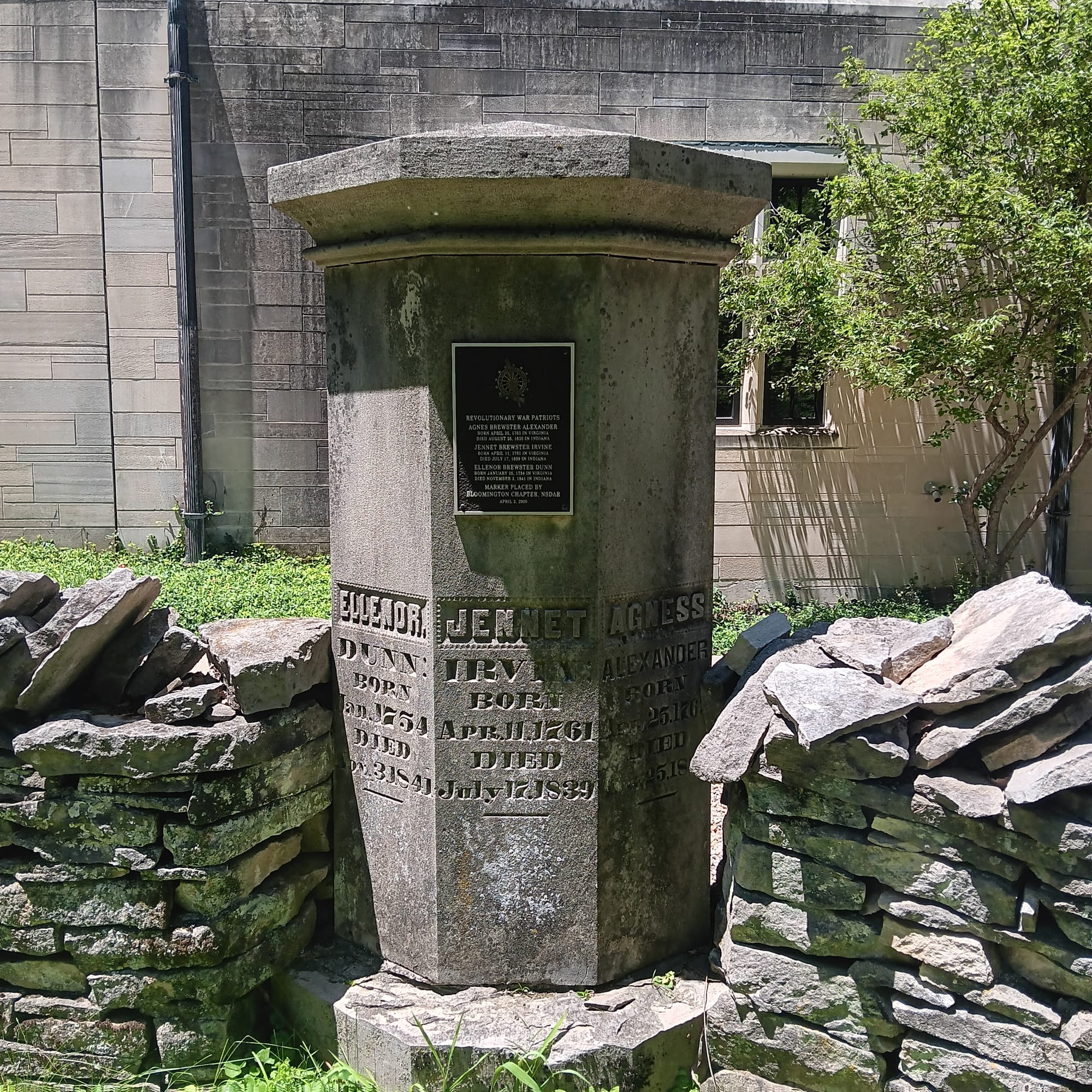 a tall columnar limestone grave marker shared by three sisters set into a limestone cobble wall. The Indiana Memorial Union building is behind