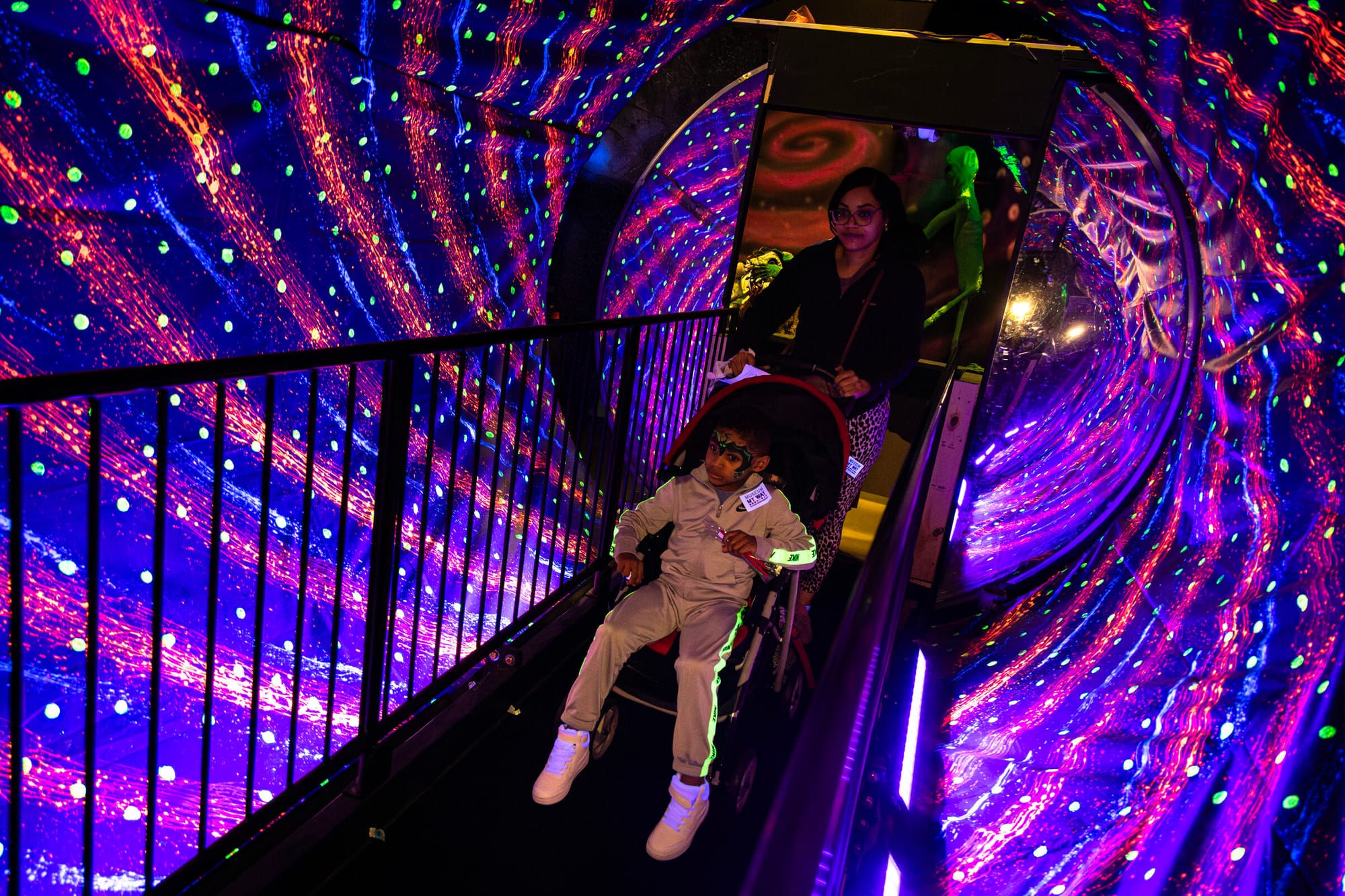 a mother pushes her child in a stroller over a walkway through a tunnel-shaped room lit up like a galaxy swirl
