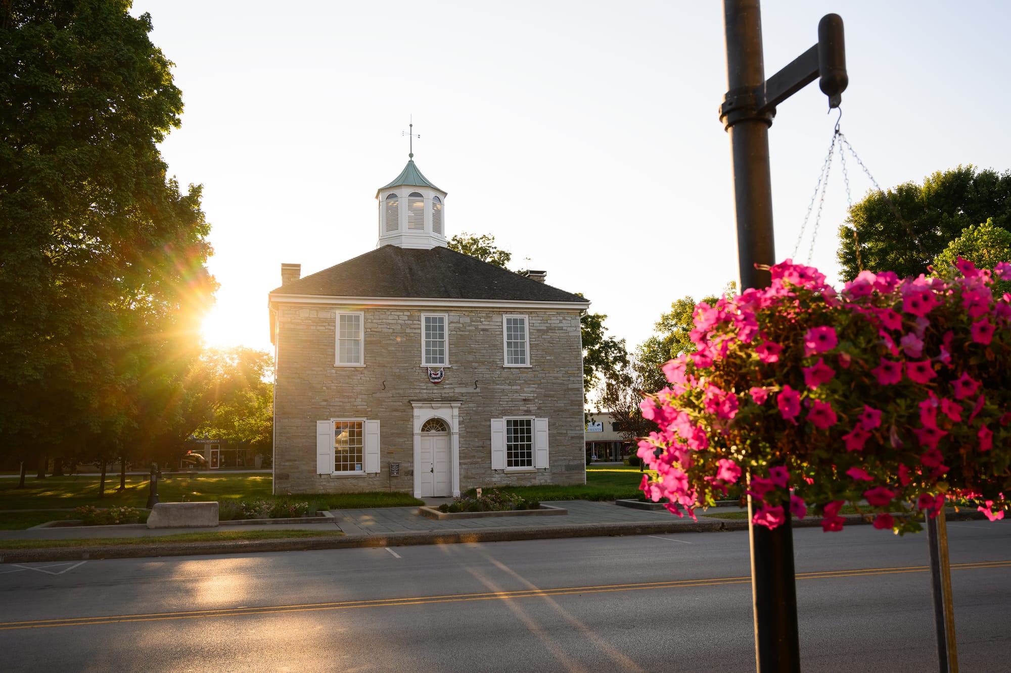 a two-story limestone building with white-paned windows. The sun sets in the background; pink flowers hang from a pole in the foreground