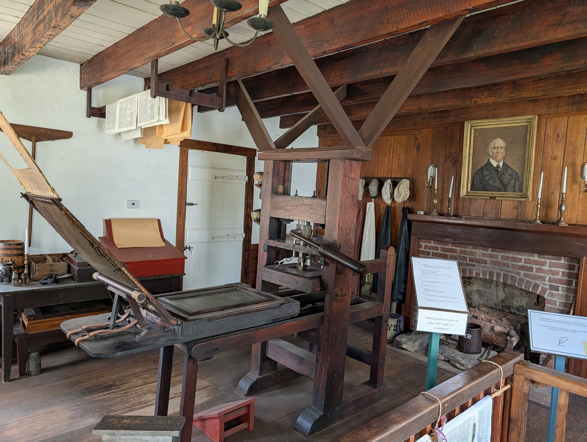 an 1800s-era wooden printing press fills much of a log cabin room. A man's portrait is mounted over a brick fireplace