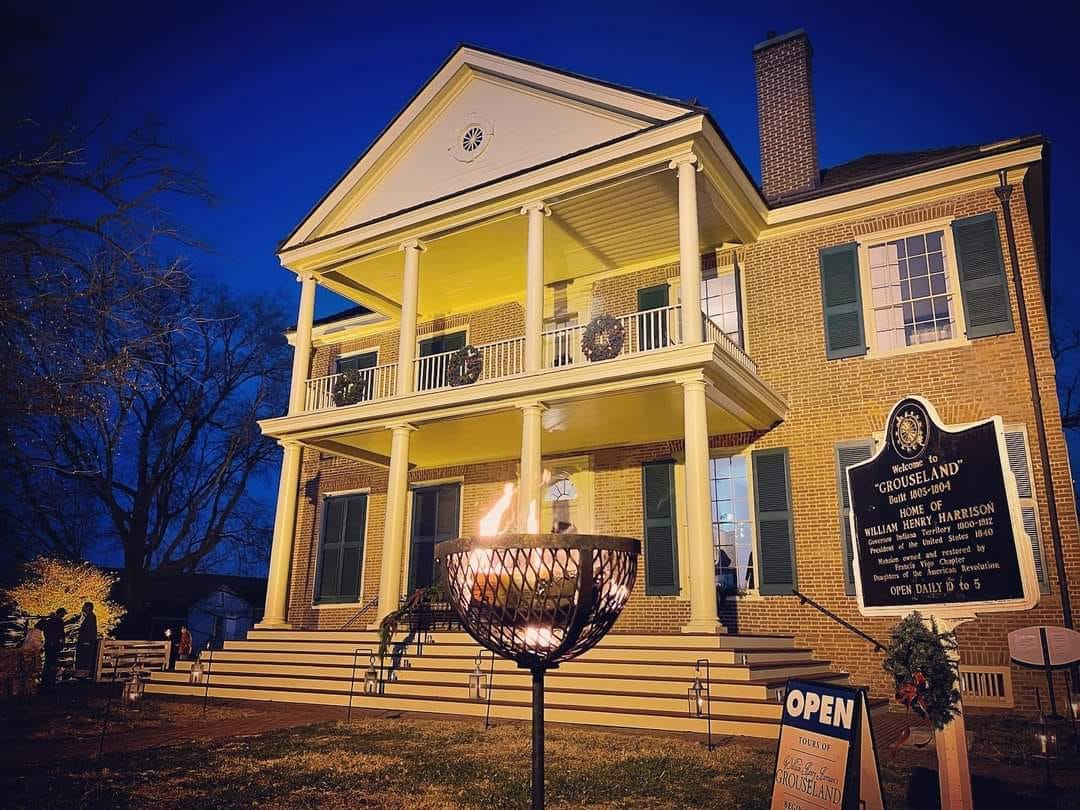 A two-story brick home with a pillared front porch and second-story balcony, each covered by a peaked roof