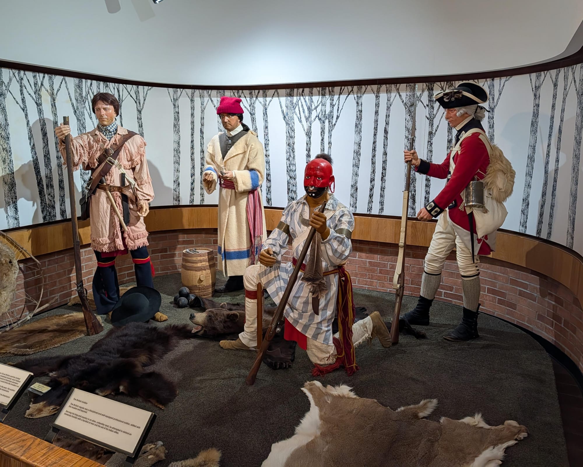 museum mannequins of an American frontiersman, a colonial-era merchant, a Native American with red and black facepaint, and a "Redcoat" British soldier posed and on display with animal-skin rugs