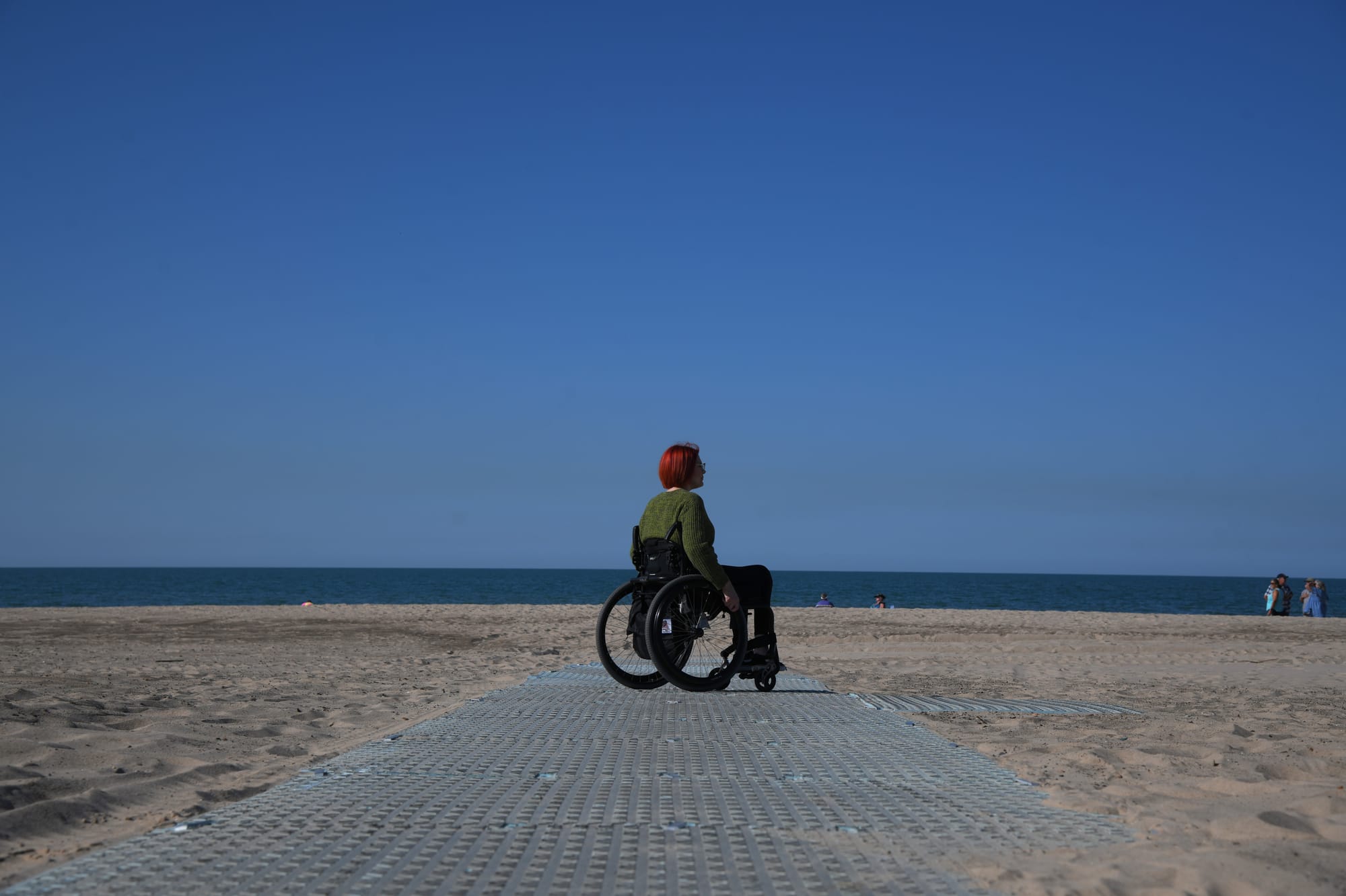 a red-haired woman wearing a green sweater sits in a wheelchair at the end of a corrugated surface on a sandy beach at Lake Michigan