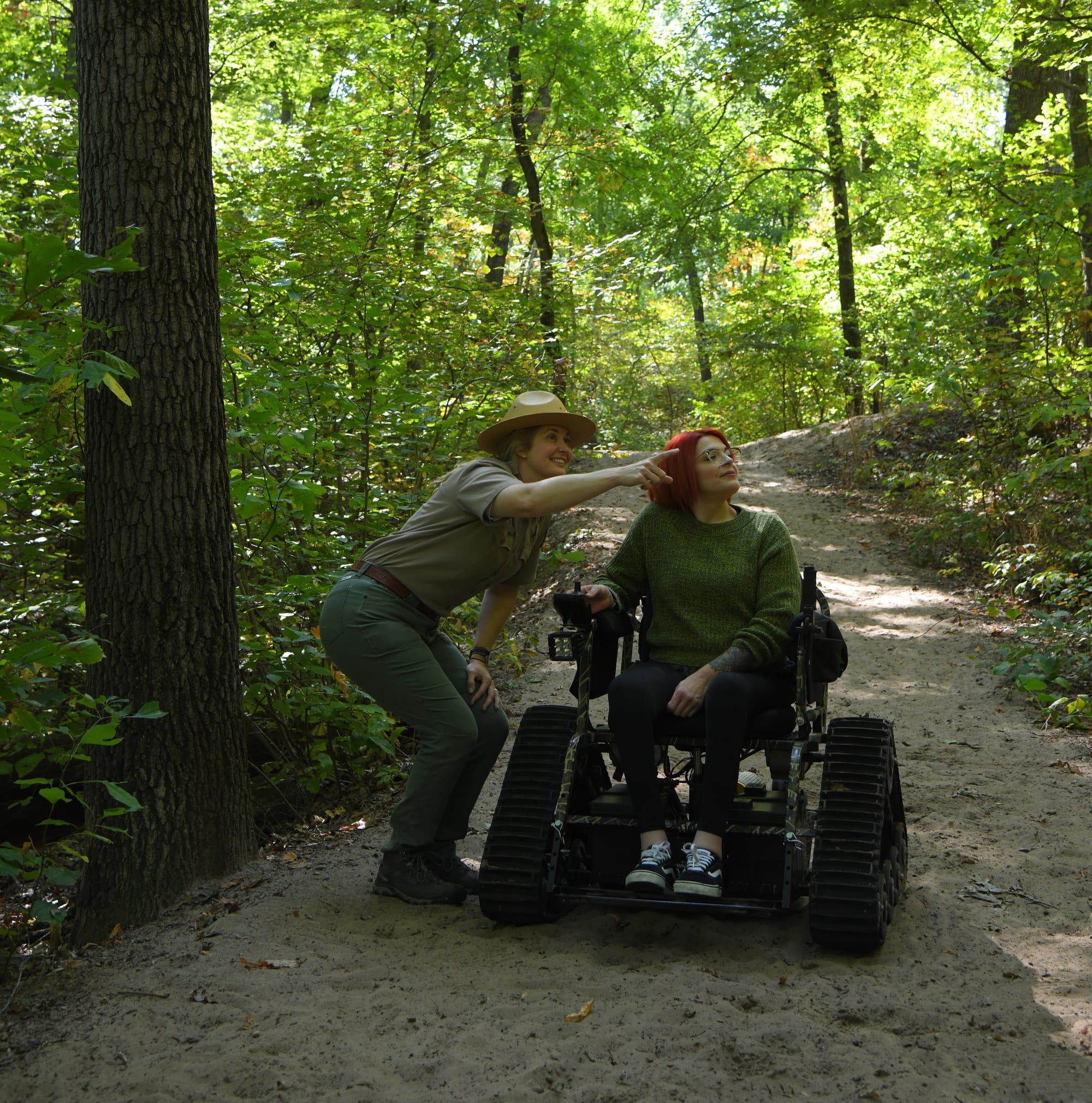 a female park ranger points into the forest; a red-haired woman in a green sweater sits in an all-terrain wheelchair on a sandy trail, looking to where the ranger points