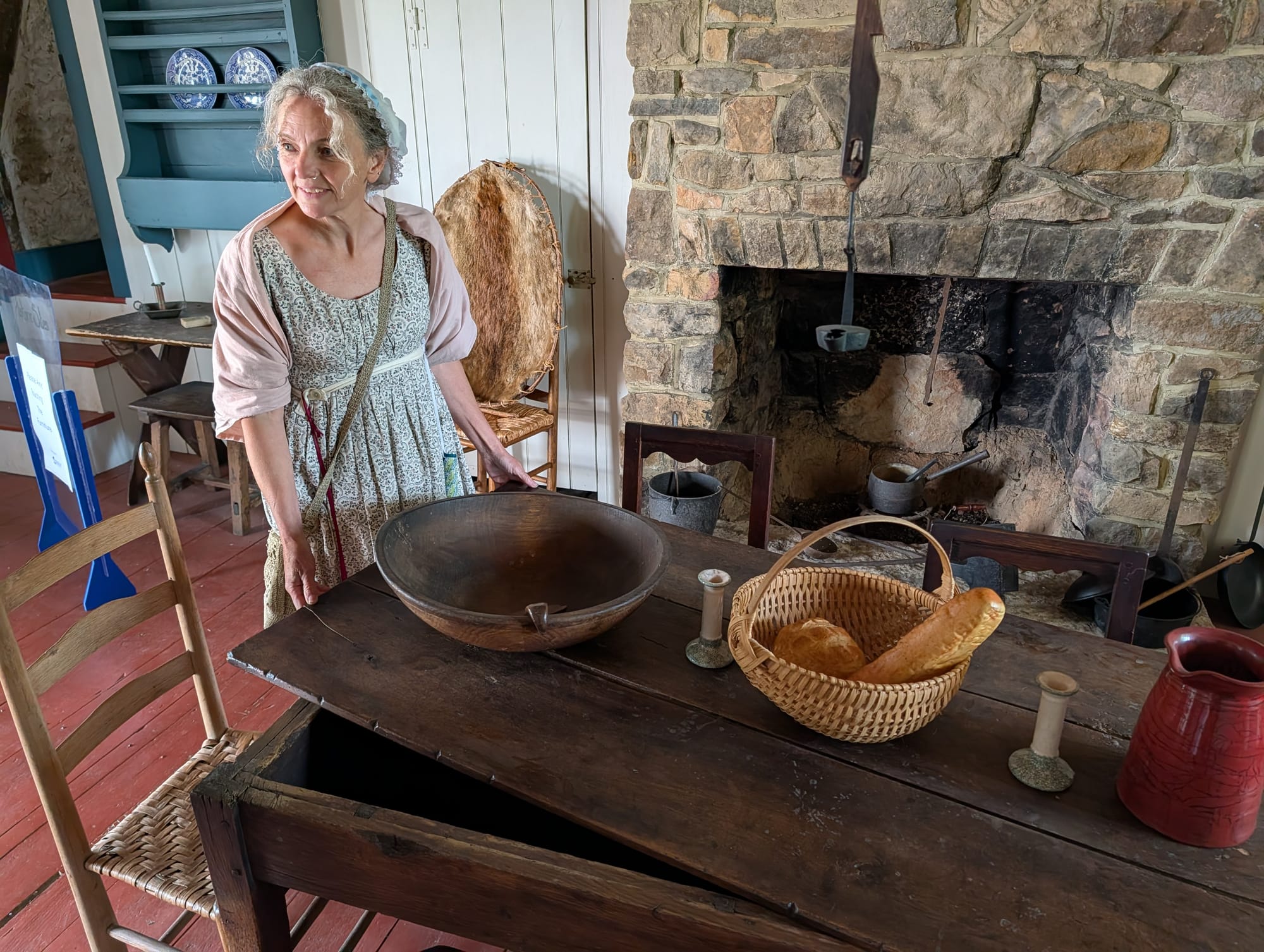 a woman in 1700s-era clothing stands at a wooden table laden with a large wooden bowl, a basket of bread, and a red ceramic pitcher