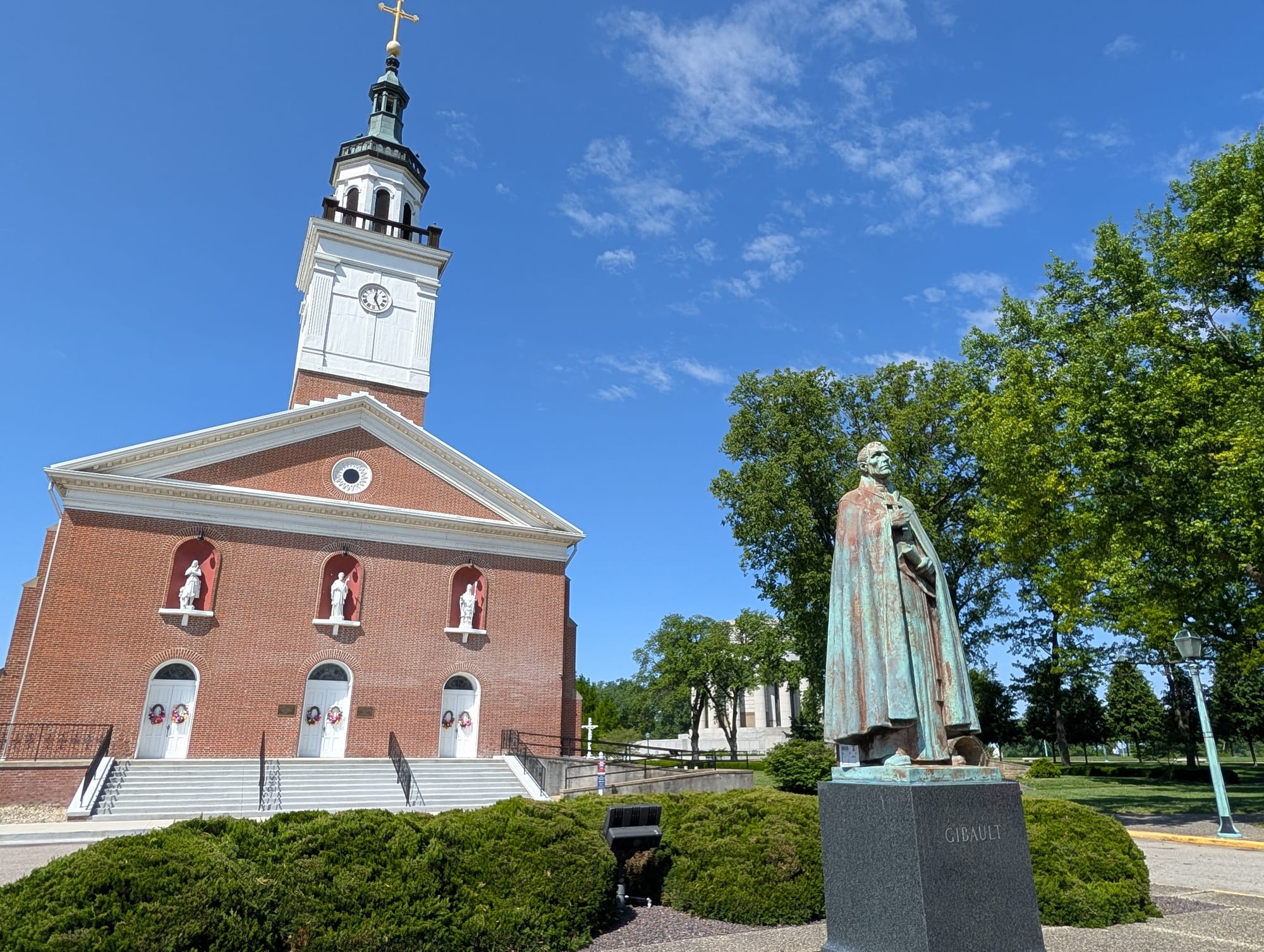 a patinaed statue of a cloaked man in front of a red brick church building with three white doors and a tall white belfry
