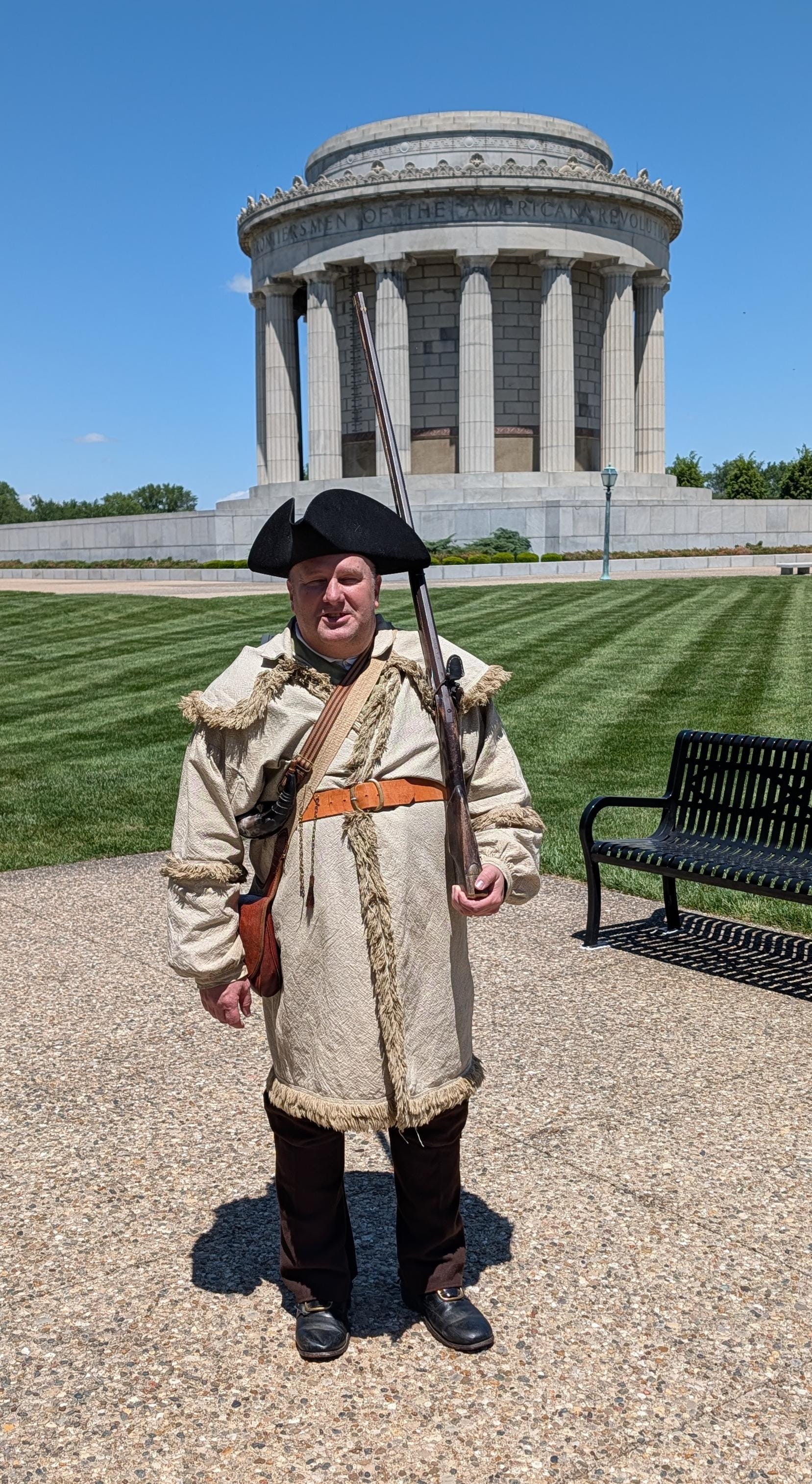a man wearing a tricorn hat, leather and fur-trimmed coat, brown pants, and black buckle shoes, stands in front of the cylindrical George Rogers Clark memorial building