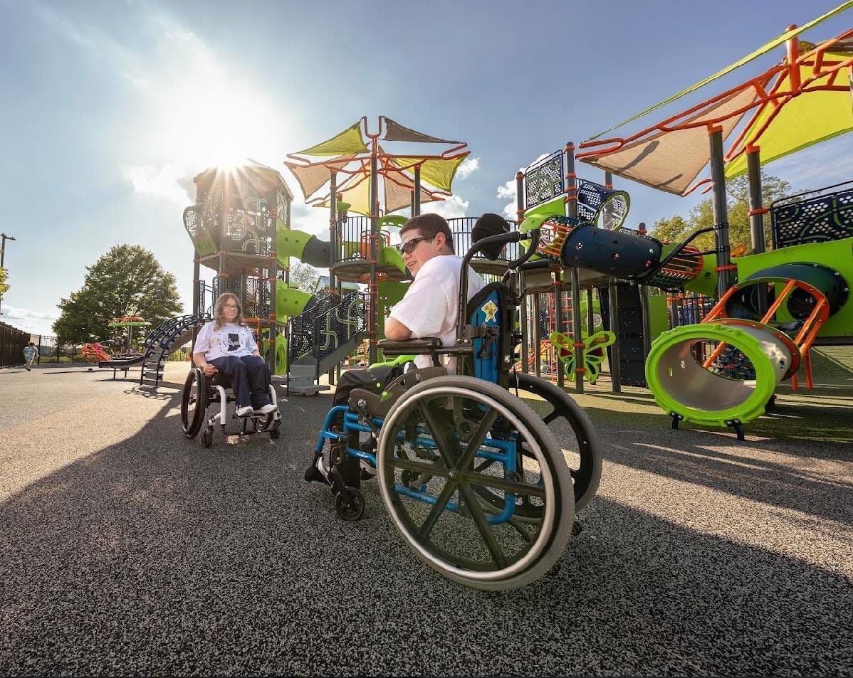 two children in wheelchairs in front of a large blue, green, and orange playground