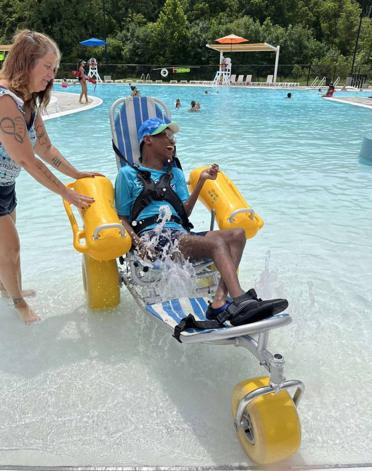 a youth enjoys the wading pool in a blue and white water wheelchair with large, yellow rubber tires and yellow flotation devices attached