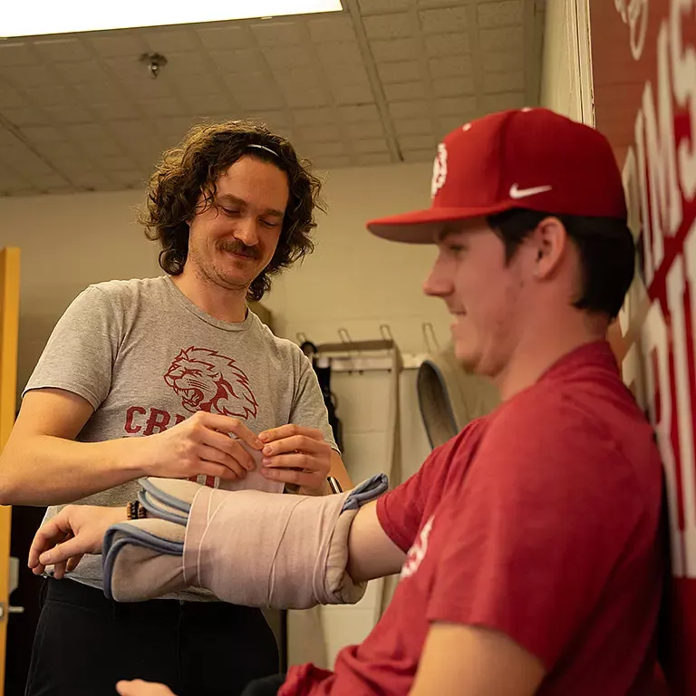 a curly-haired man wearing a grey t-shirt wraps medical tape around the arm of a young athlete wearing a red baseball cap and a red t-shirt