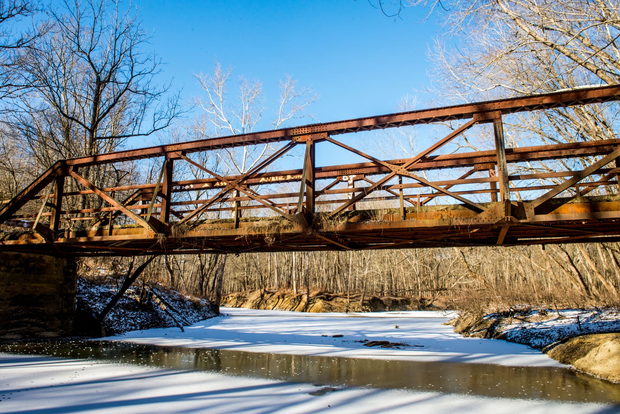 a rust-colored iron bridge spans an icy creek in winter
