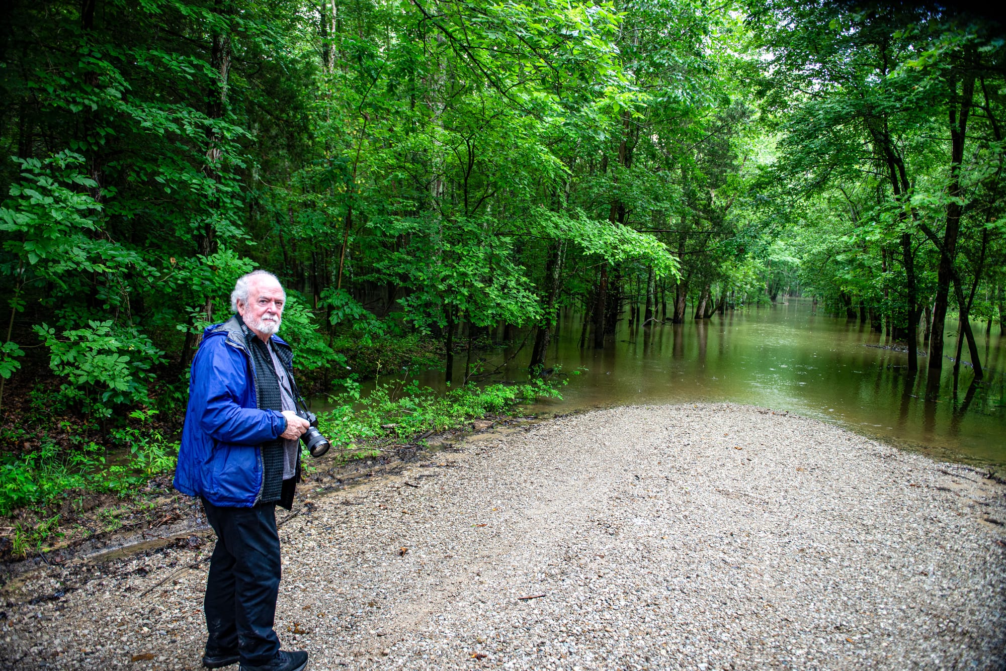 an elderly man in a blue coat holds a camera near the end of a gravel road which becomes submerged in water a short distance away