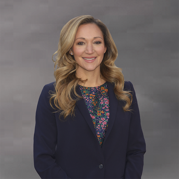 a woman with curling brown-blonde hair, wearing a blue blazer over a floral shirt