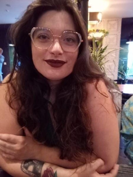 a young woman with long brown hair, large glasses, dark lipstick, and a forearm tattoo