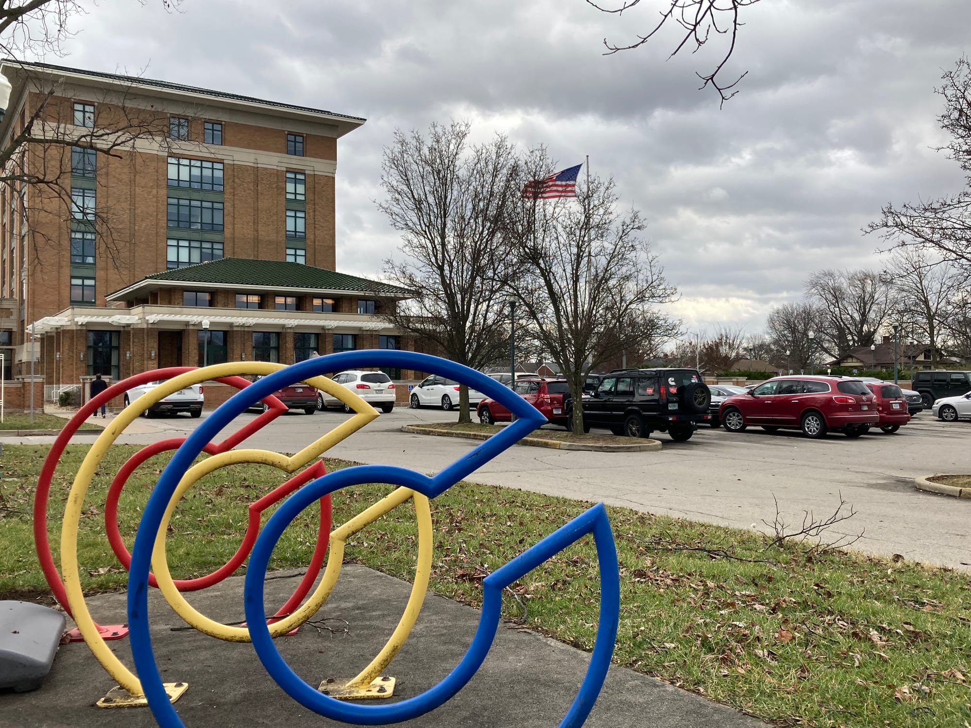 three metal bicycle racks shaped like the letter "C" painted one each blue, yellow, and red. A parking lot, American flag, and brown brick hospital building in the background