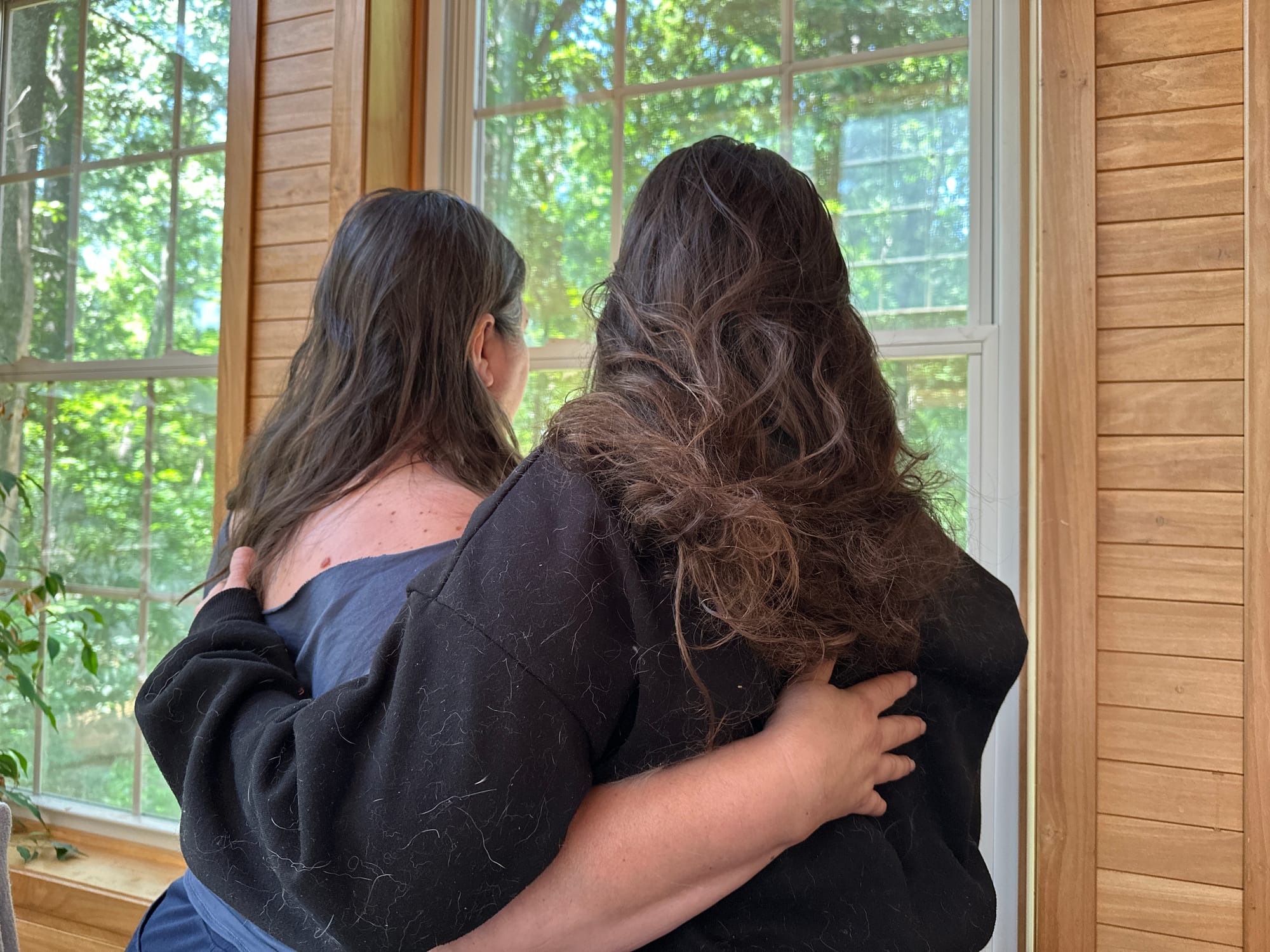 two women with long brown hair stand arm-in-arm facing a window, their backs turned to the camera