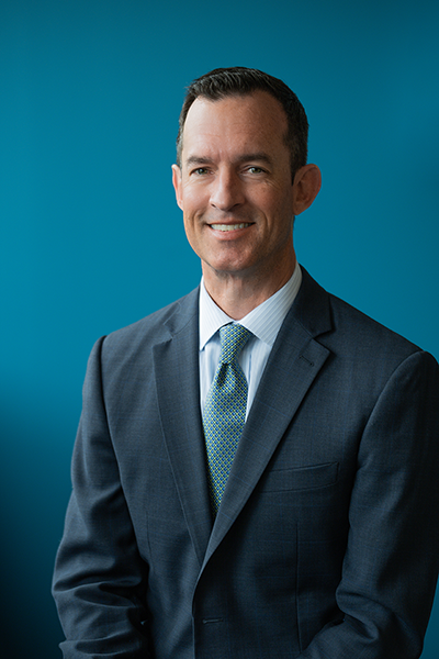 an austere yet friendly looking man in a grey jacket, light blue shirt, and blue tie, against a blue backdrop
