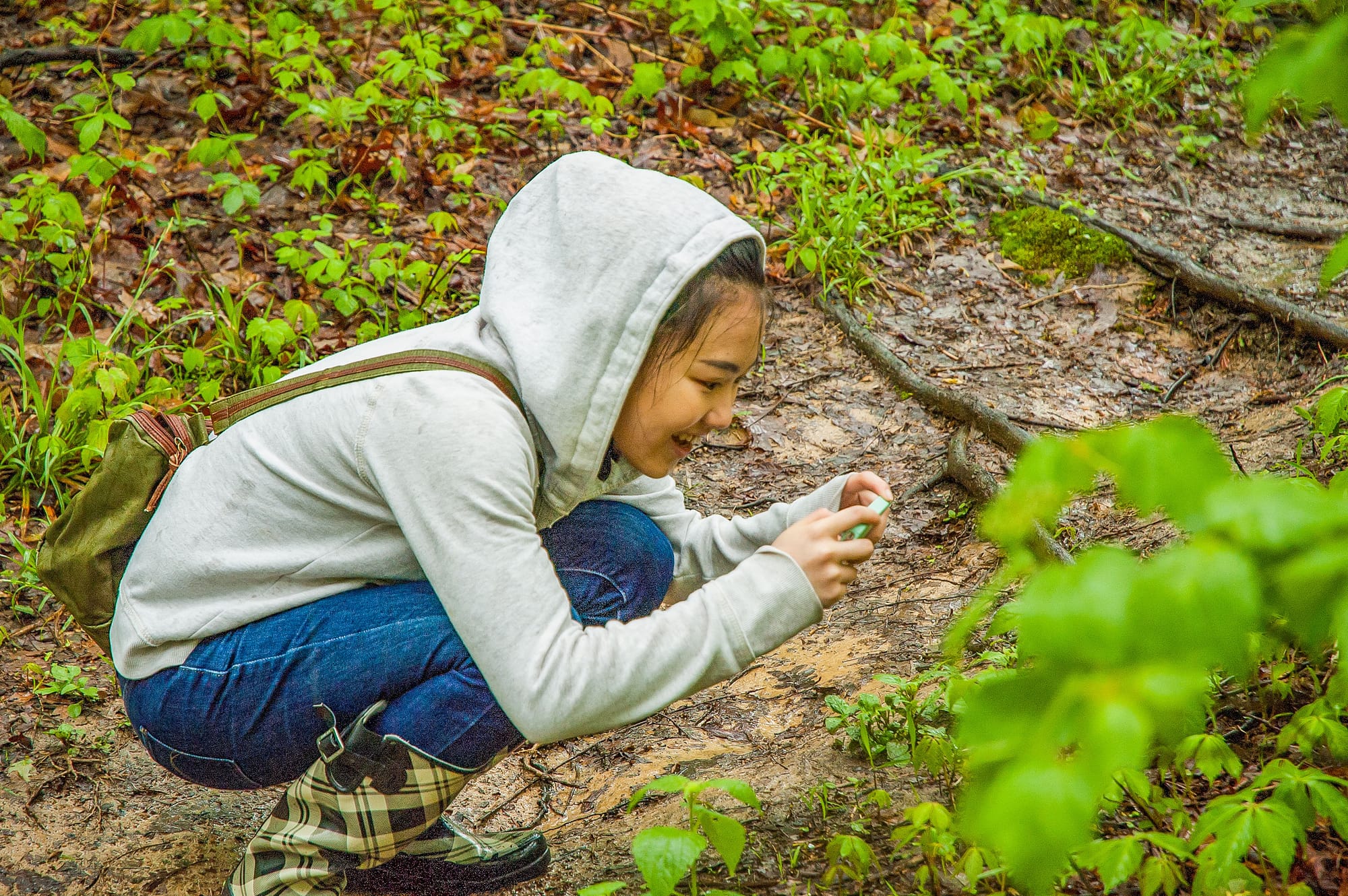 a young woman wearing rain boots, blue jeans, and an ash grey hooded sweatshirt, crouches to photograph flora along a trail with her smartphone
