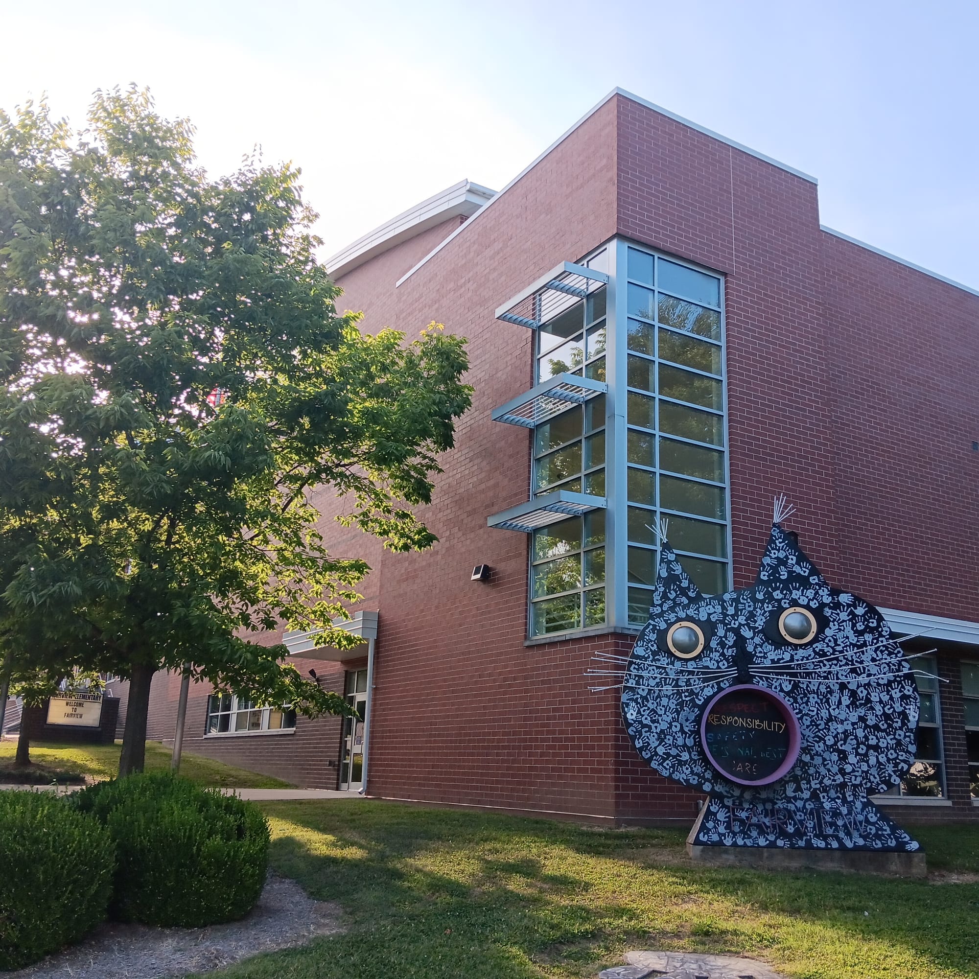 a tall brick school building with curving roof architecture. Trees and bushes shade the building's south wall. A large, cartoonish metal bust of a cat, painted with children's handprints, decorates the exterior