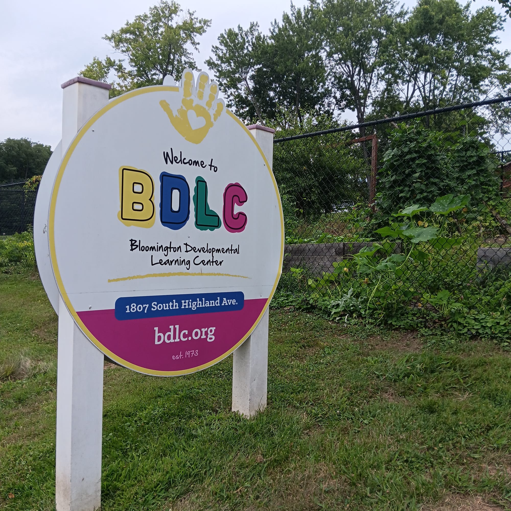 a circular white and pink sign with a yellow handprint painted at the top. It reads: "Welcome to BDLC Bloomington Developmental Learning Center, 1807 South Highland Ave., bdlc.org, est. 1973"
