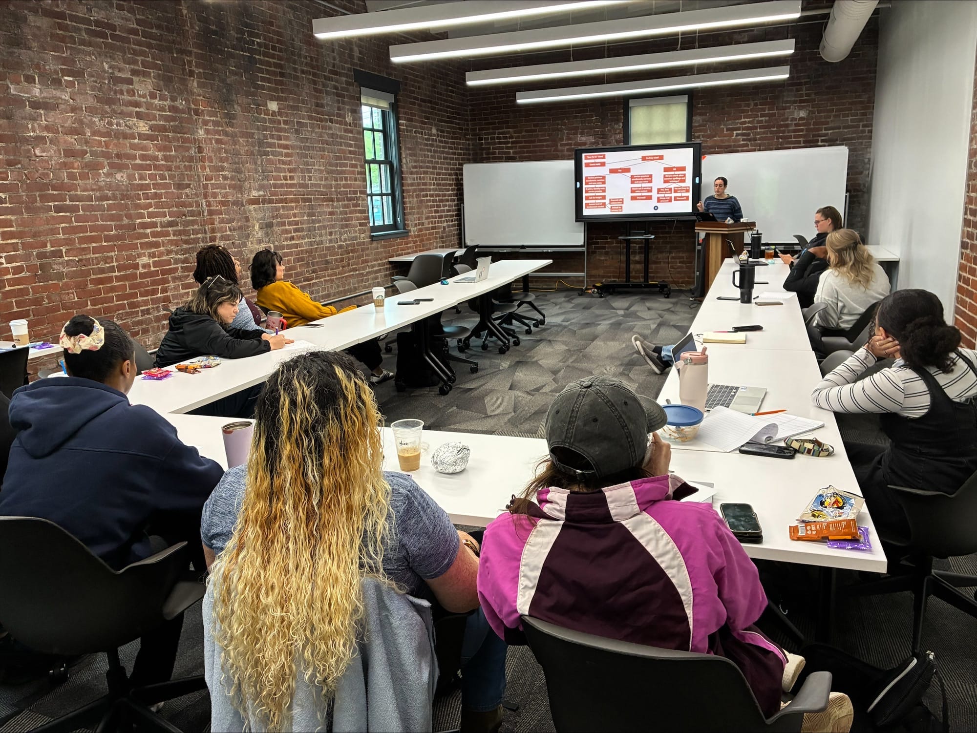 Ten individuals sit around a U-shape of rectangular white tables inside a brick-walled room; a woman at a podium presents a PowerPoint