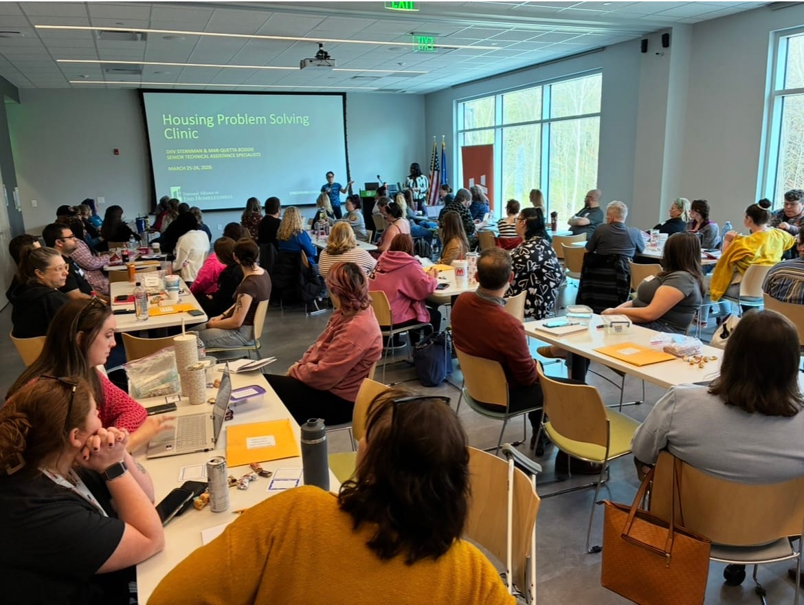 a room full of attendees sitting at scattered tables; presenters lead the seminar with a PowerPoint